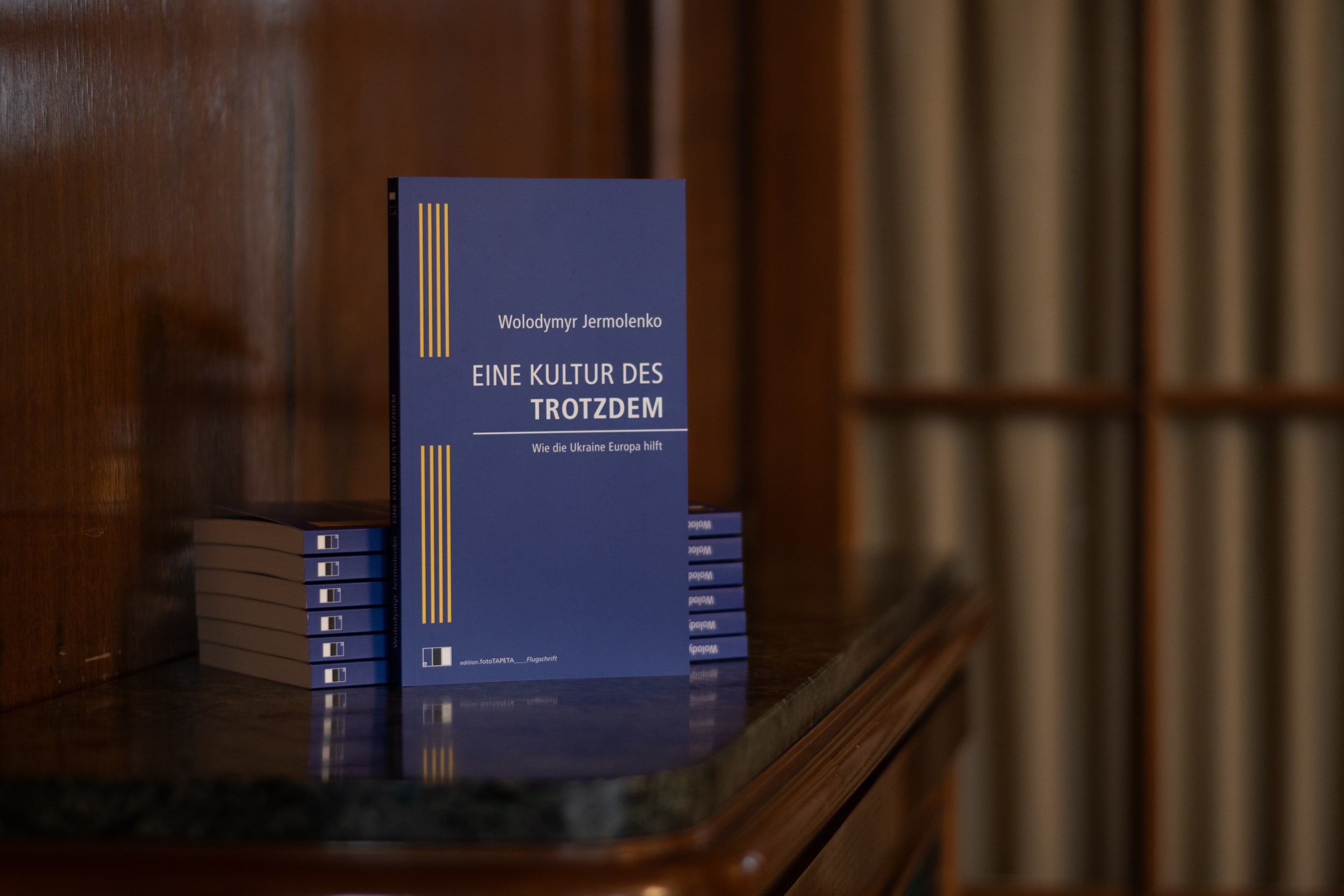A blue book titled "Eine Kultur Des Trotdzem" with yellow lines on the left side is on a wooden bookshelf, with other books in the background.