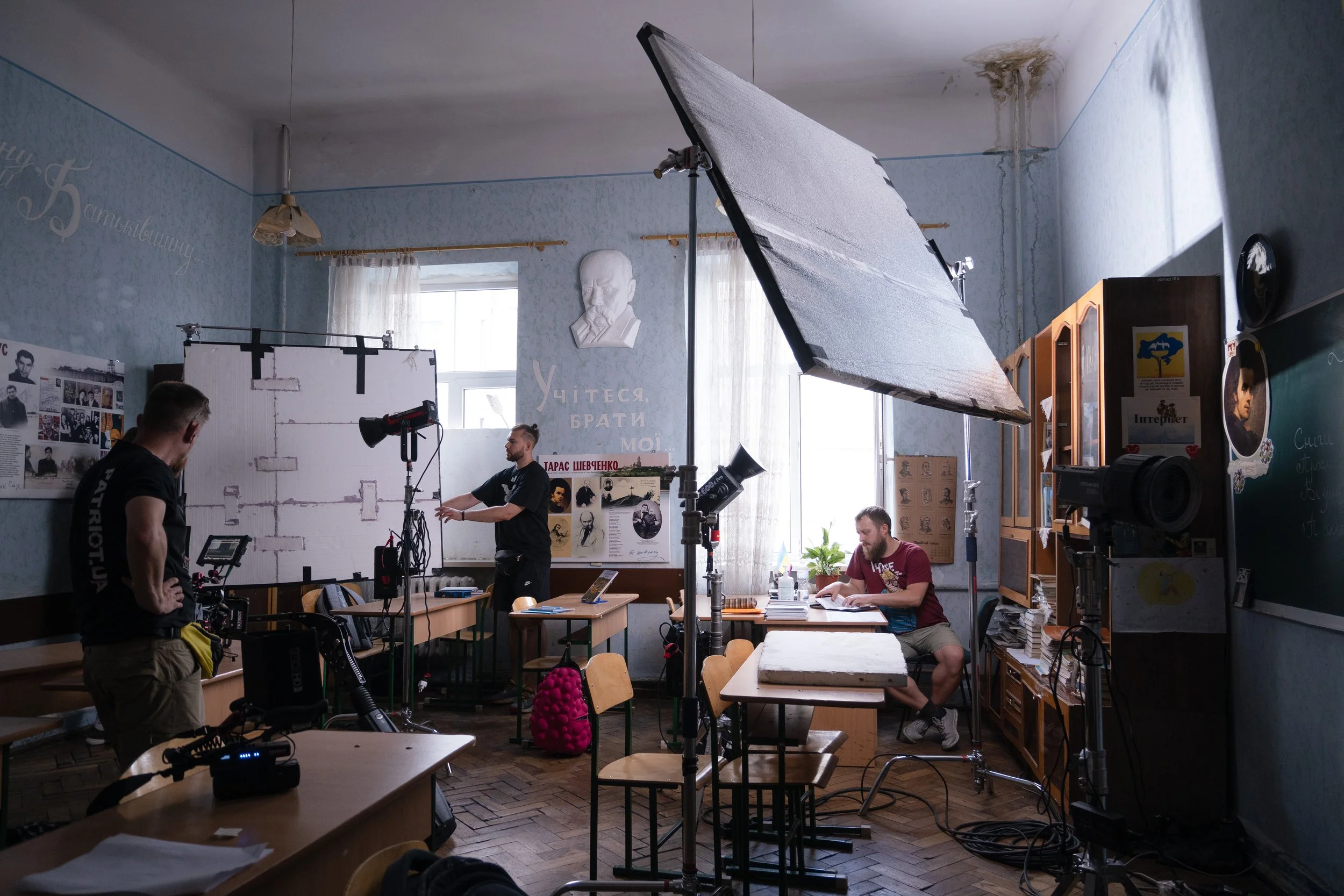 A filming or photography studio set up inside a room with multiple lighting equipment, cameras, and crew members preparing for a shoot. There are tables, chairs, and a chalkboard with some posters and pictures on the walls.