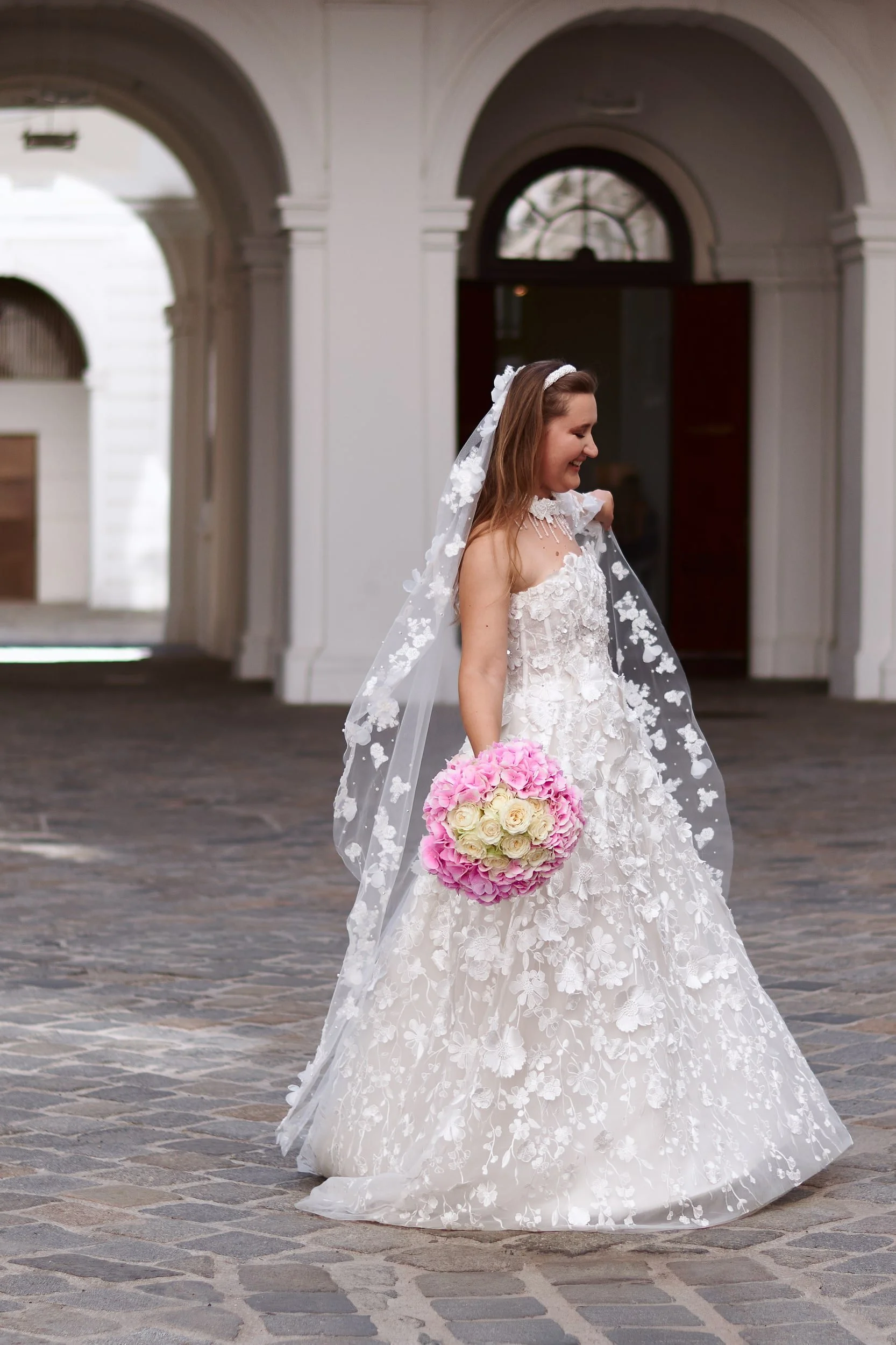 A smiling bride in a white lace wedding gown holding a bouquet of pink and white roses, standing on a cobblestone street in front of a white building with arches.