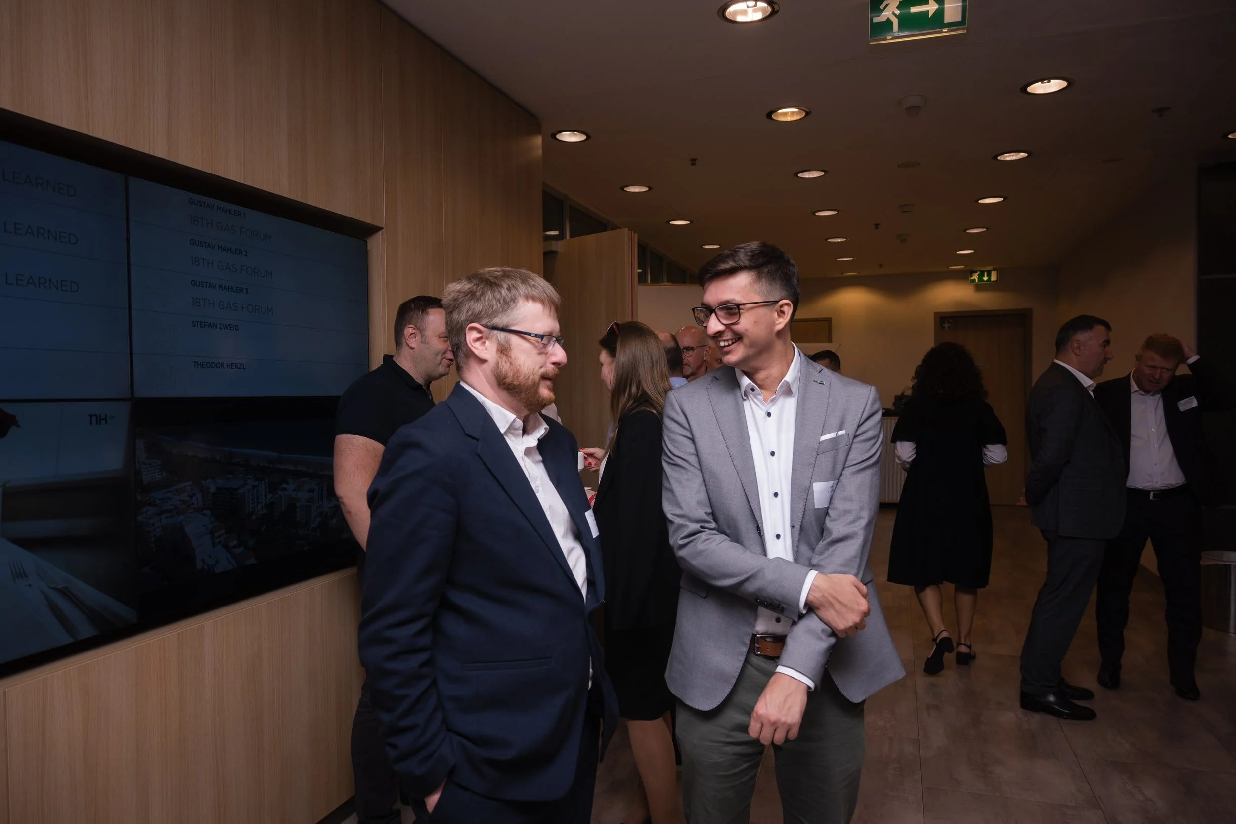 Two men in business suits are talking and smiling at a professional networking event, with other attendees visible in the background.