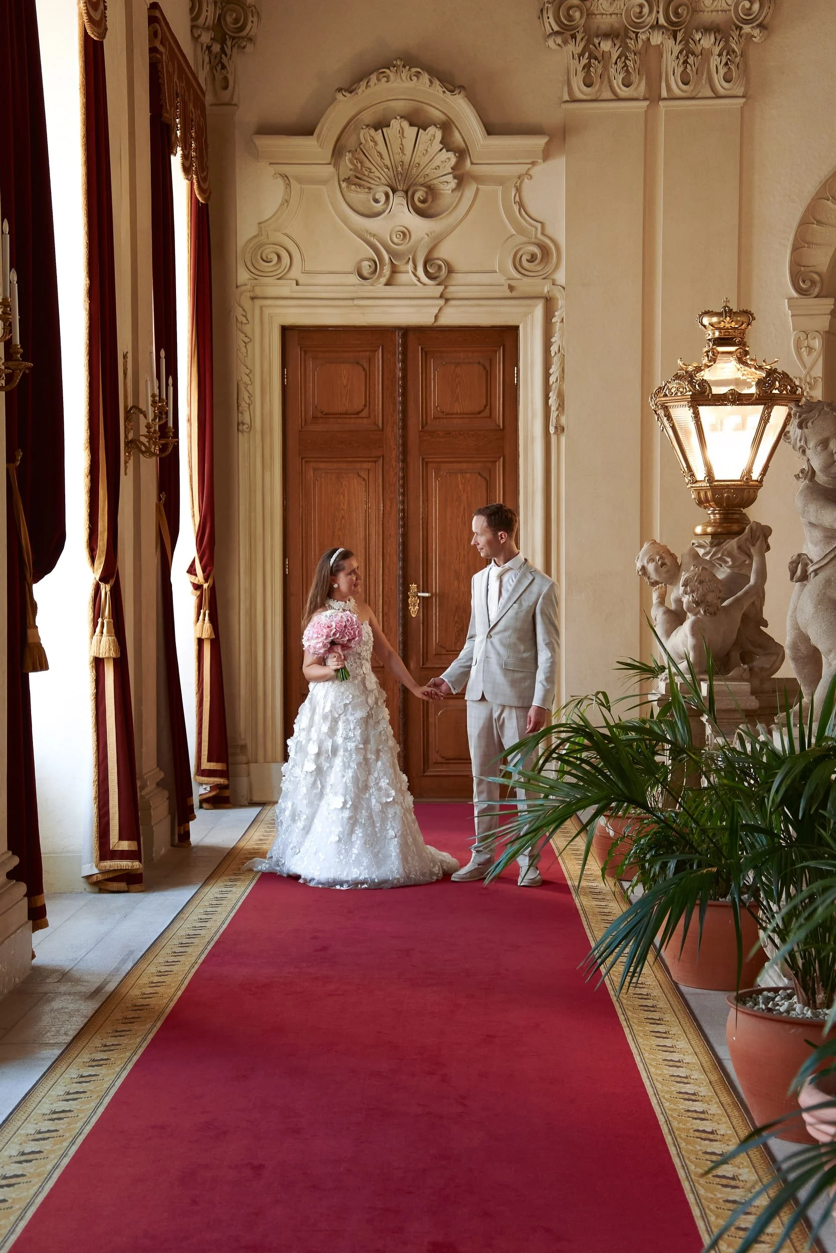 A bride and groom holding hands in a grand hall with ornate decor, wooden doors, a red carpet, and decorative statues and lamps.