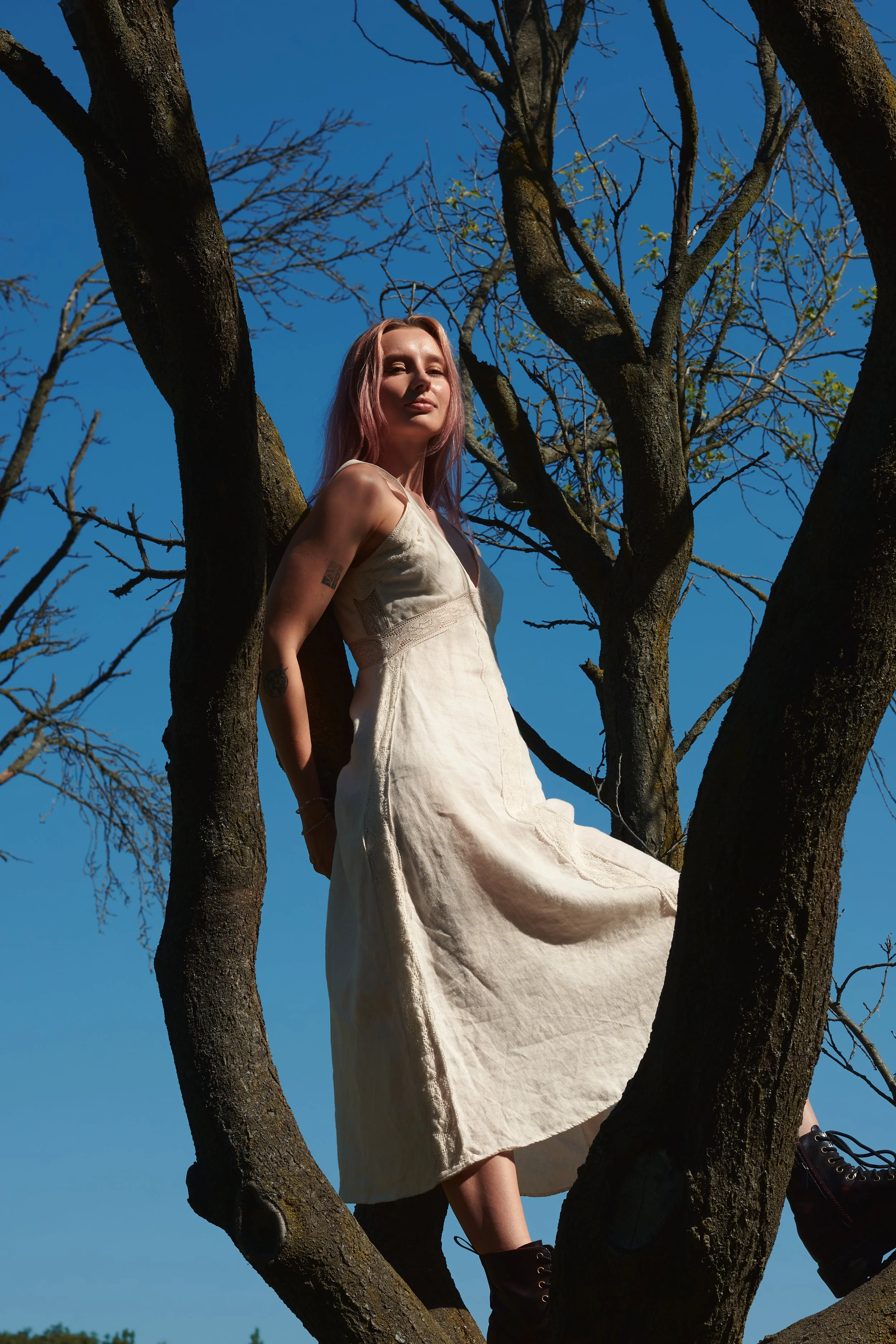A woman with pink hair stands on tree branches against a clear blue sky, wearing a light-colored dress and black boots.