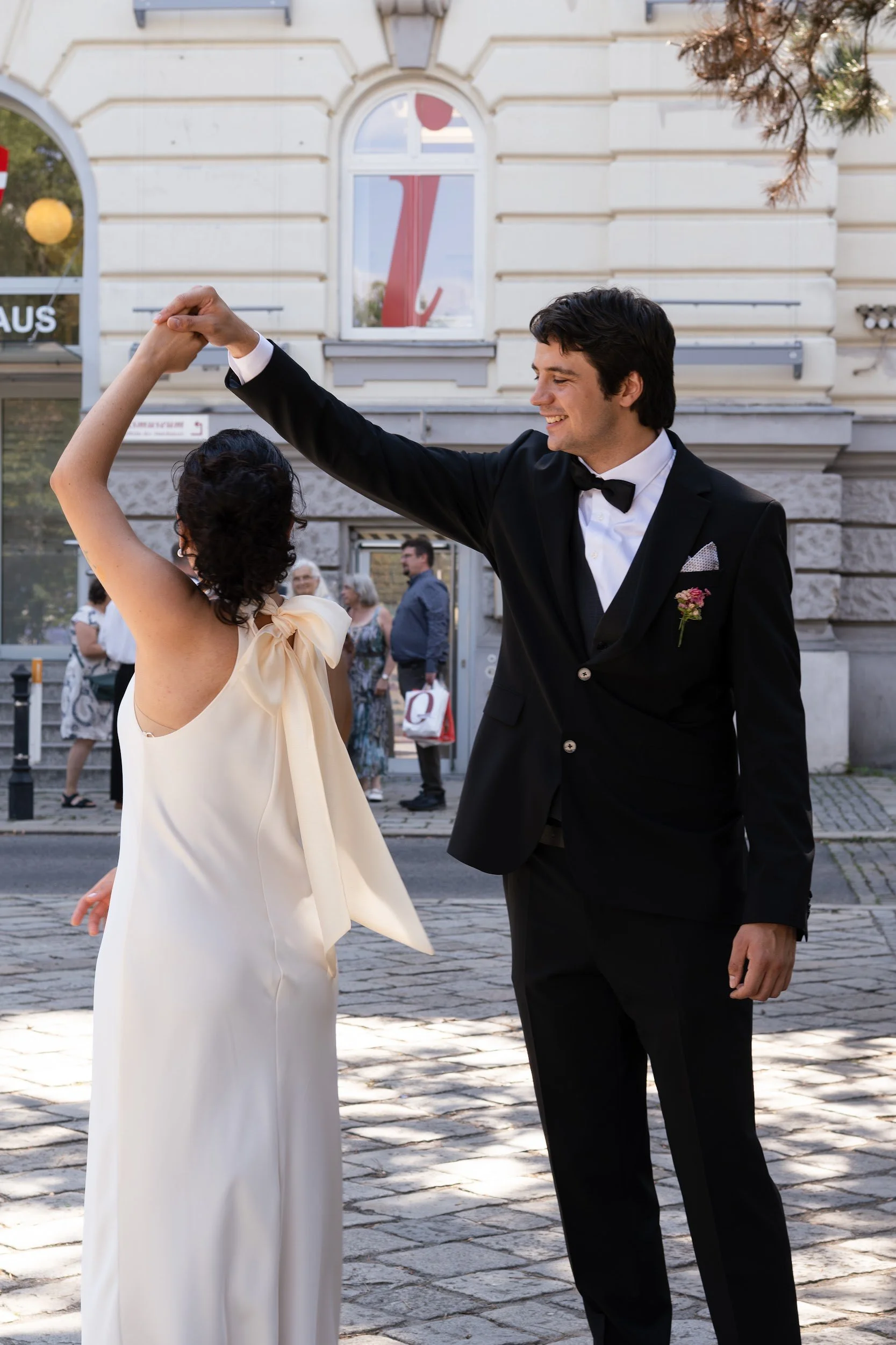 A man and woman in formal attire dancing outdoors in front of a historic building, with onlookers in the background.