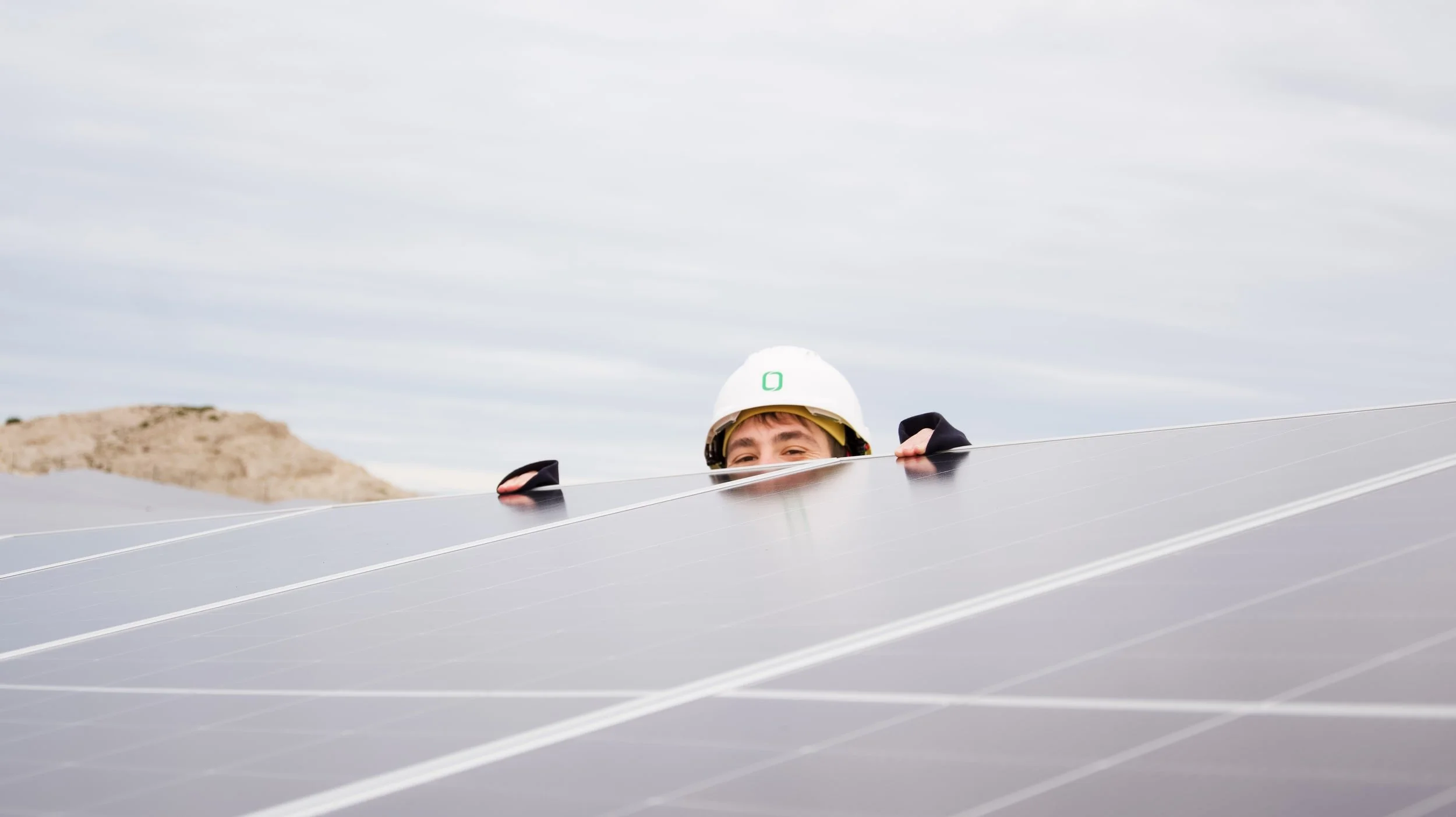 Person wearing a white safety helmet peeking over a large solar panel installation outdoors.