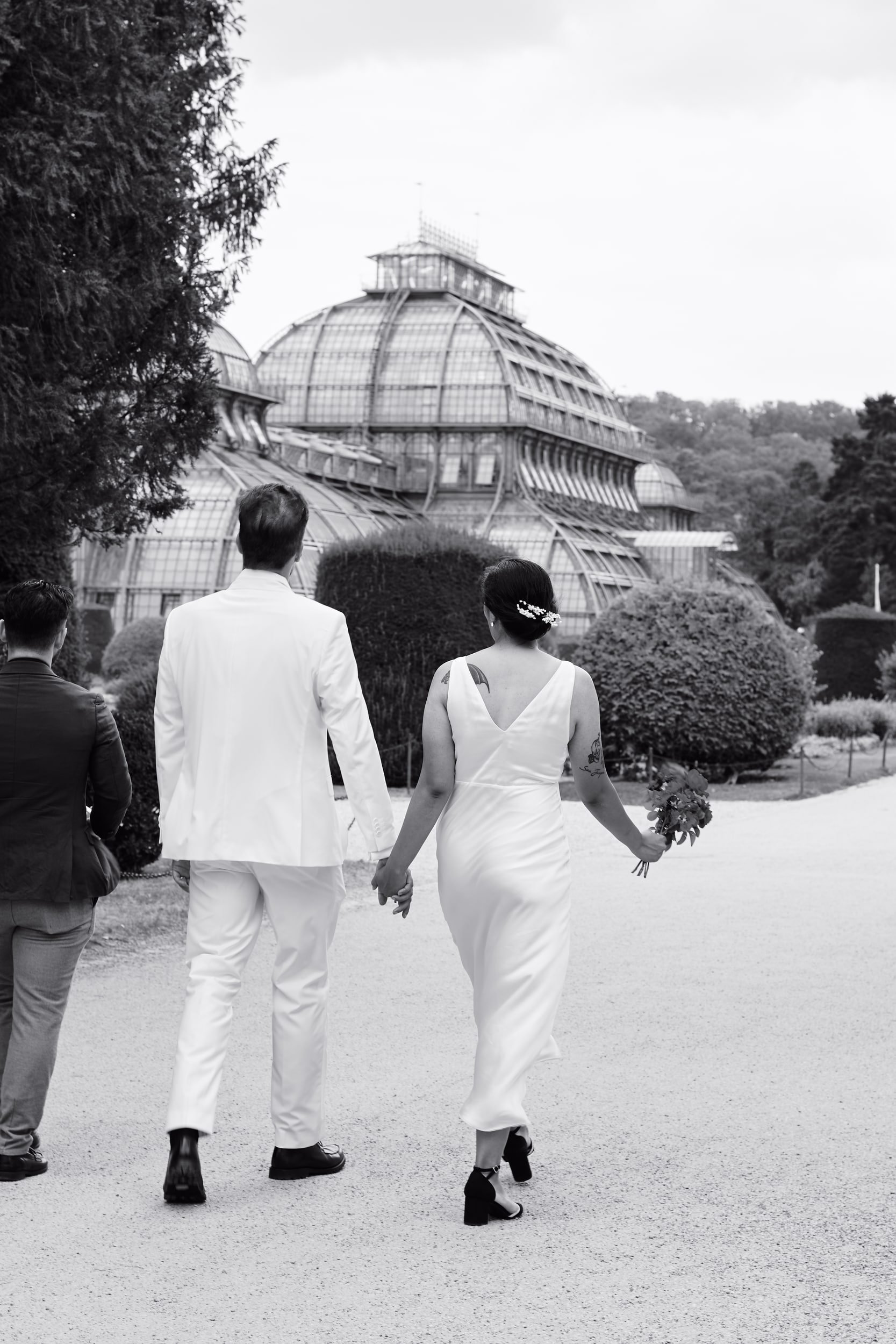 A black and white photo of a couple walking hand in hand outdoors, with a glass building and trees in the background.