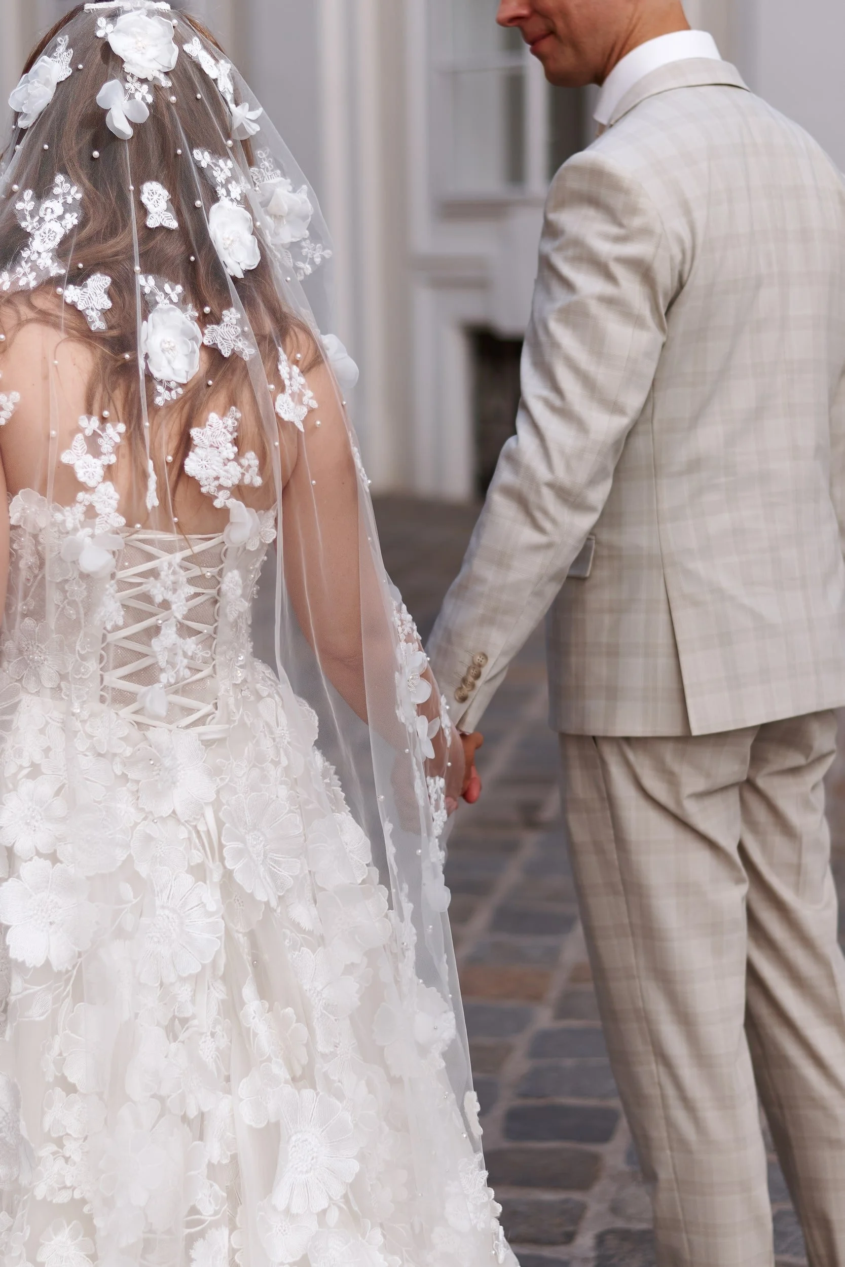 A bride and groom holding hands during their wedding ceremony. The bride wears a detailed white wedding dress with floral lace and a veil adorned with 3D floral appliqués and pearls. The groom wears a light checkered suit.