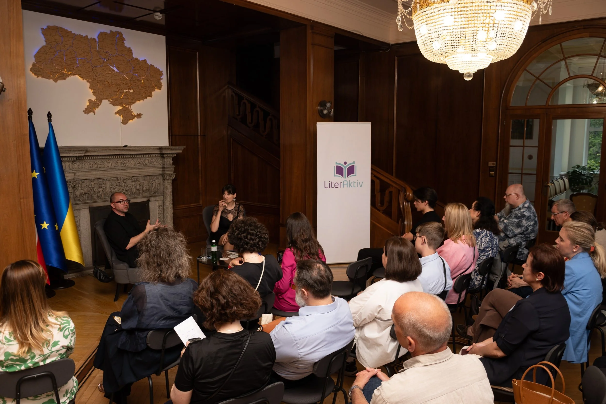 A group of people attending a presentation or panel discussion in a formal room with wooden paneling, a chandelier, and a decorative fireplace. Two individuals are seated at the front, one man and one woman, with the woman drinking from a cup. The au