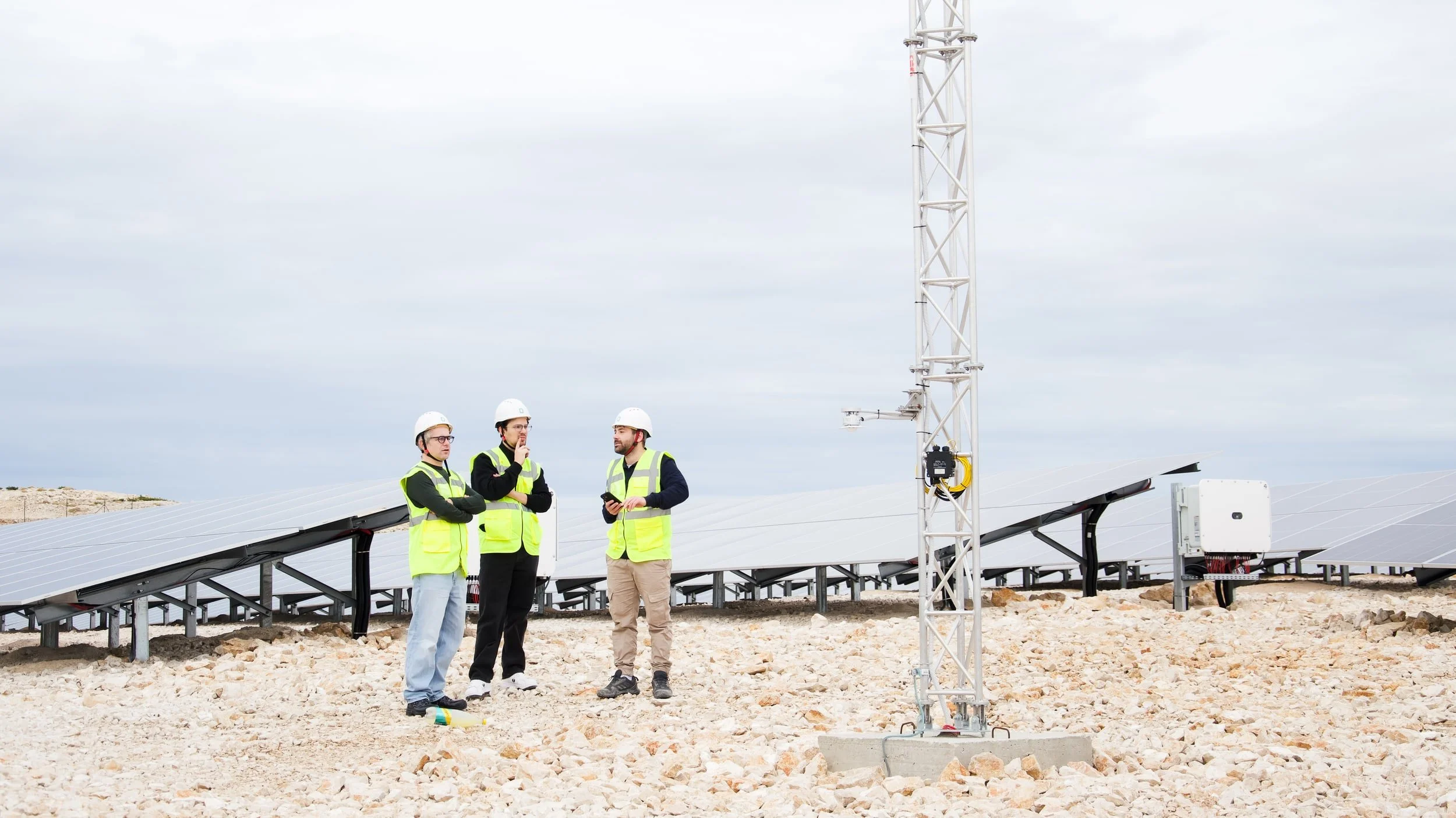 Three workers in safety vests and helmets standing near solar panels and a tall communication tower during cloudy weather.