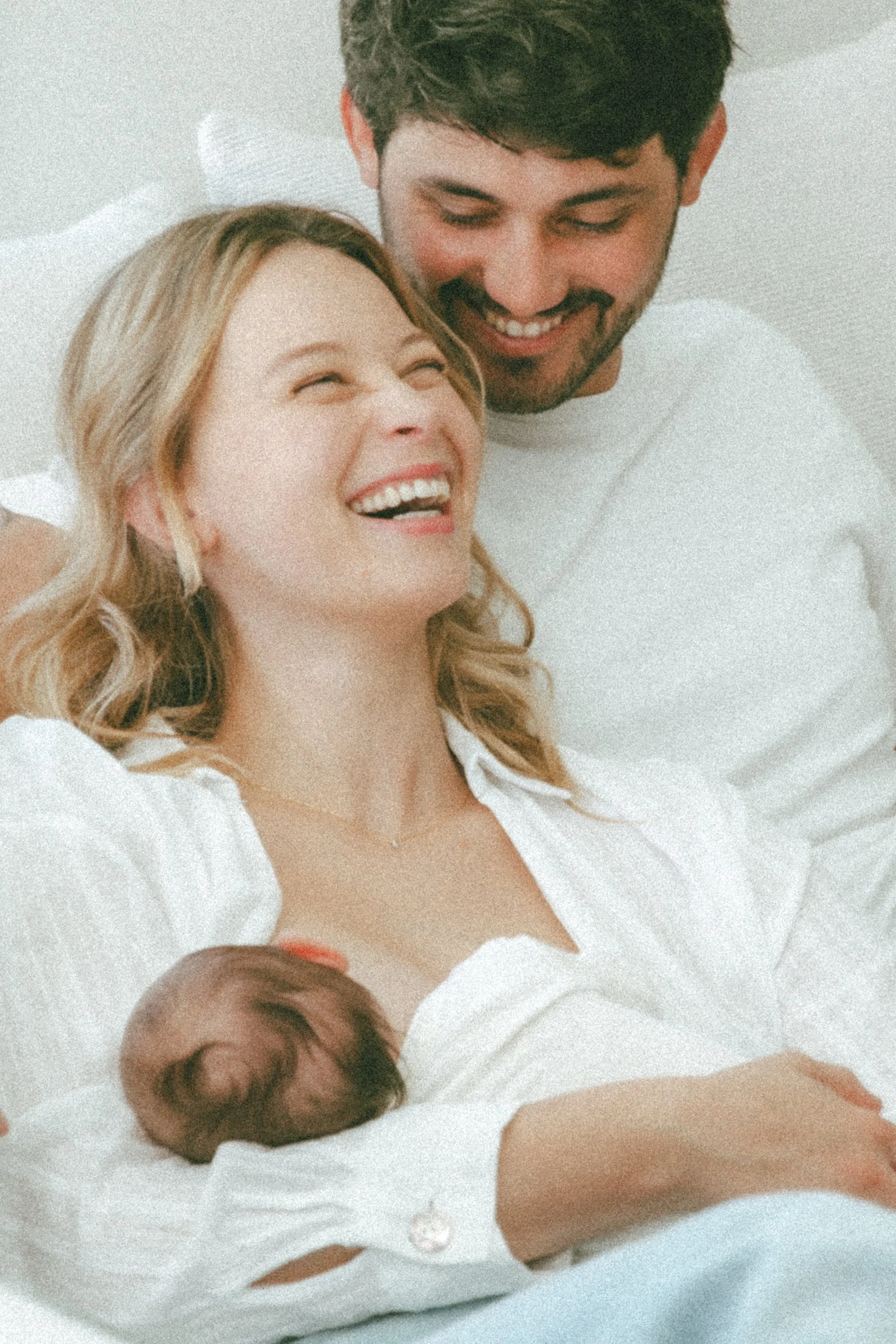 Une femme et un homme souriants, allongés ensemble dans un lit blanc, partageant un moment de bonheur, avec un bébé près d'eux.