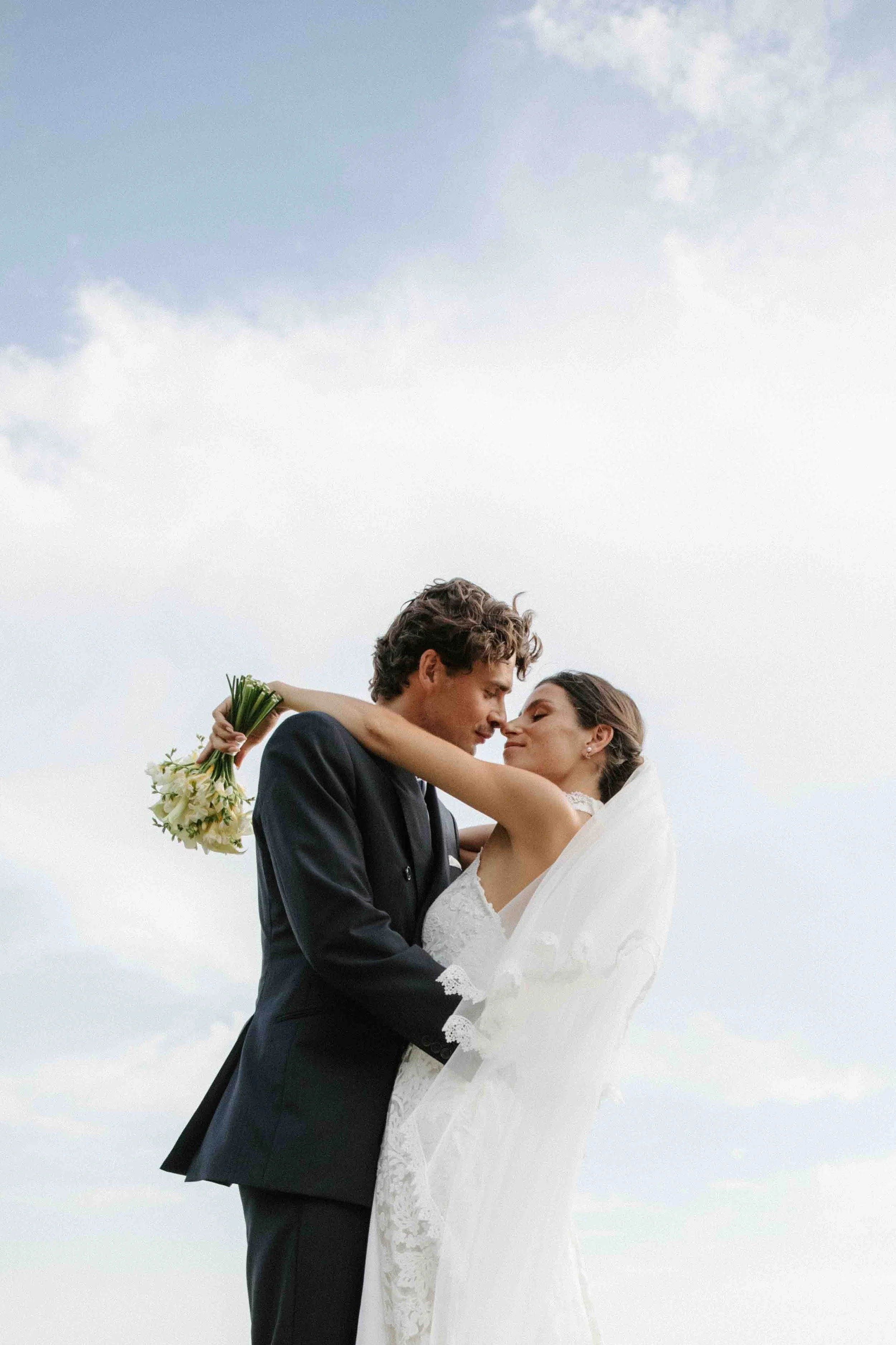 Un couple de mariés s'embrassant lors de leur mariage en plein air, la mariée portant une robe blanche et le marié un costume noir, avec un ciel nuageux en arrière-plan.