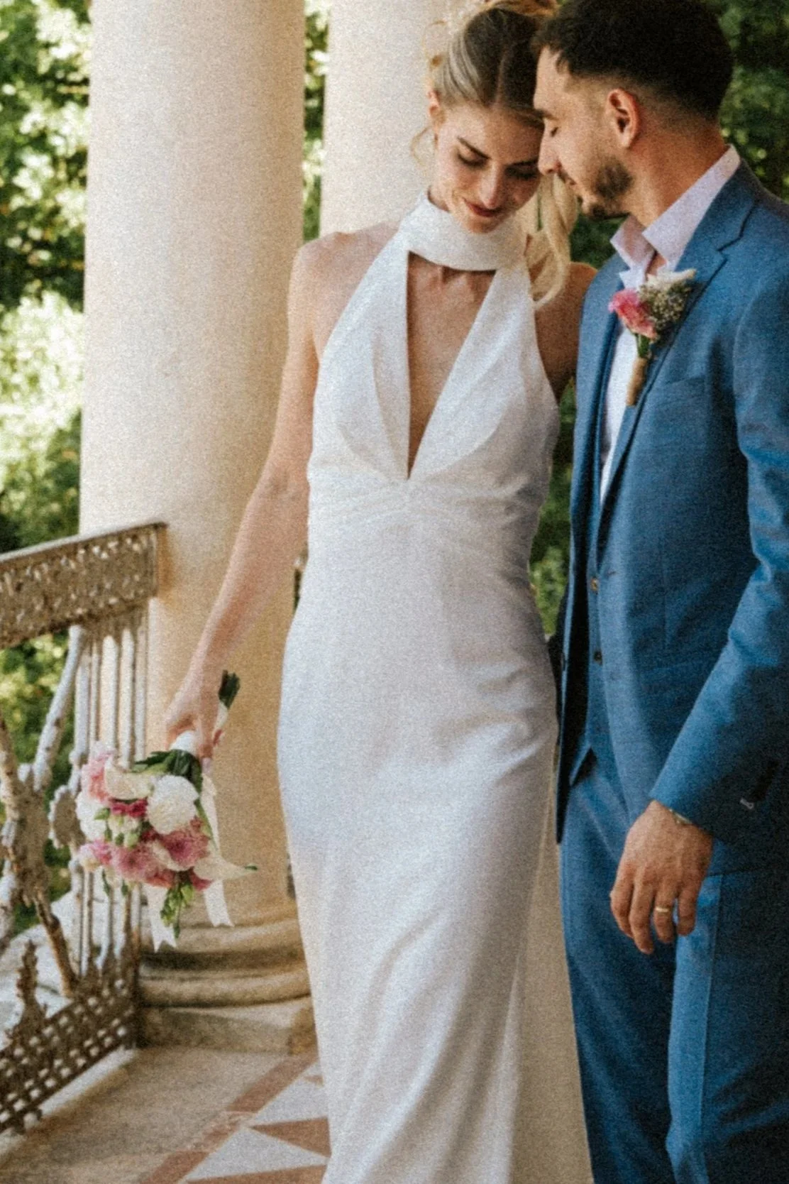 Un couple en mariage, la femme en robe blanche et le homme en costume bleu, posant près d'une colonne sur un balcon avec un bouquet de fleurs.