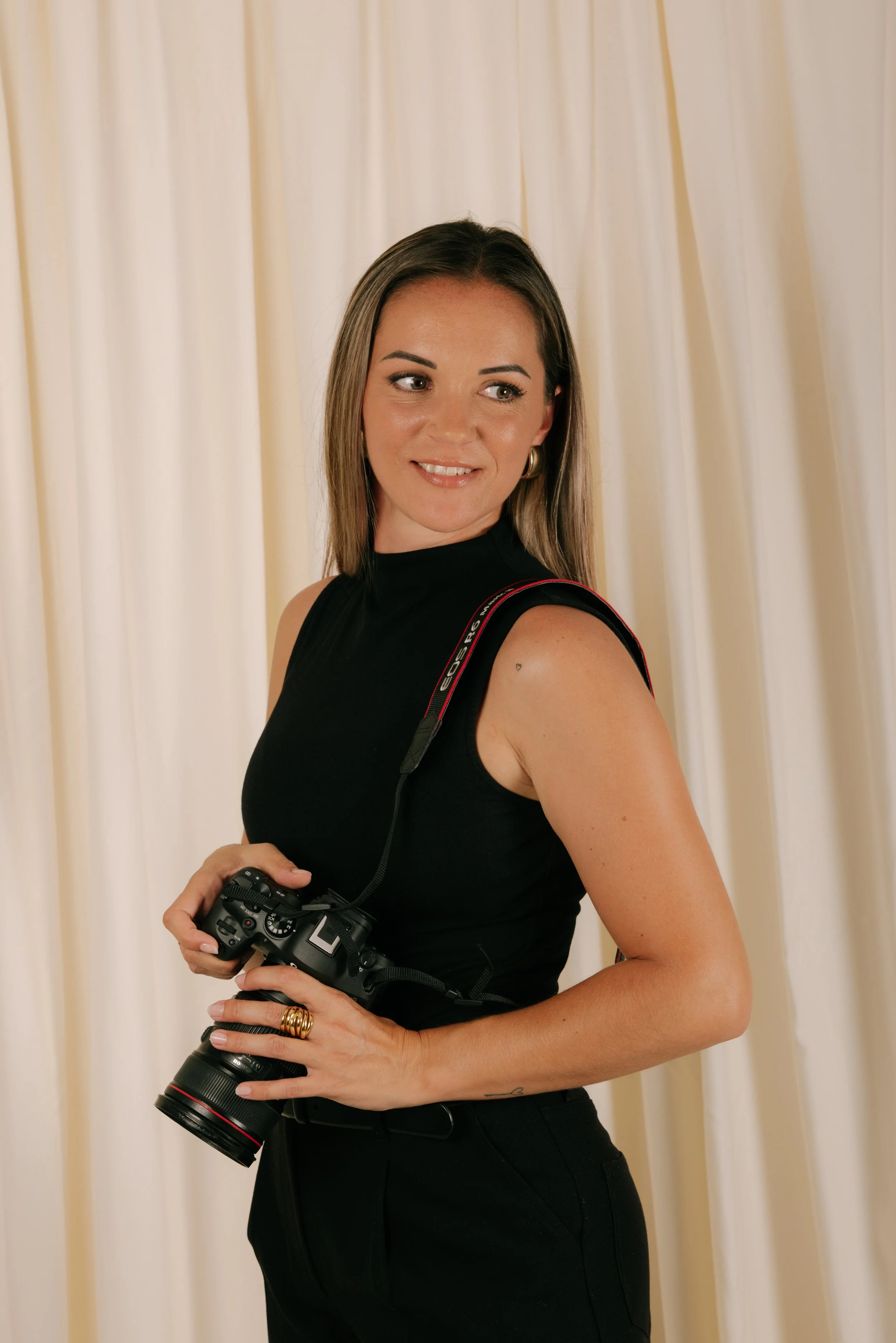 Une femme avec des cheveux bruns, portant un haut noir, tenant un appareil photo professionnel, se tient devant un rideau beige clair.