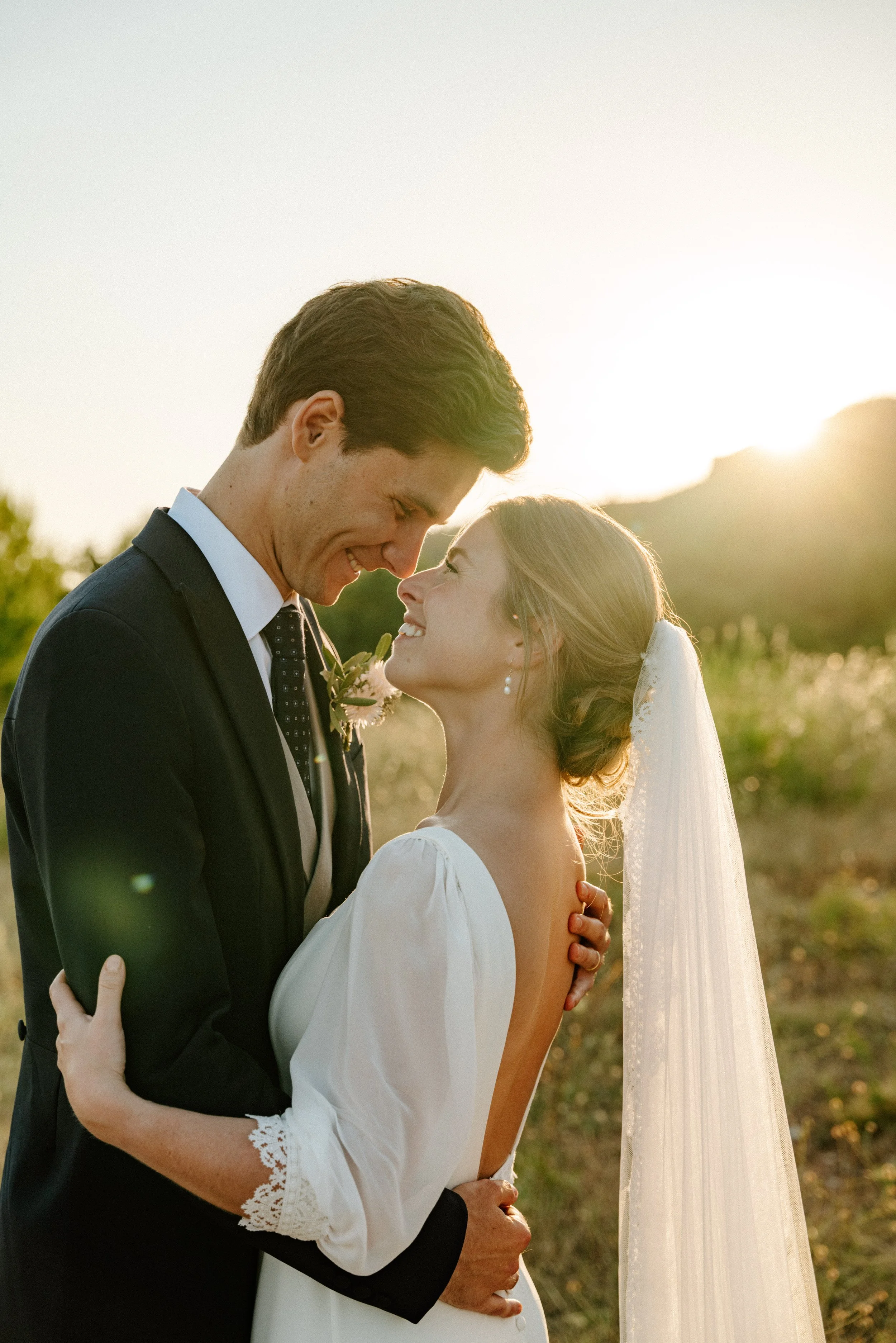 Un couple de jeunes mariés se regarde tendrement lors d'une séance photo en plein air au coucher du soleil, dans un paysage verdoyant.