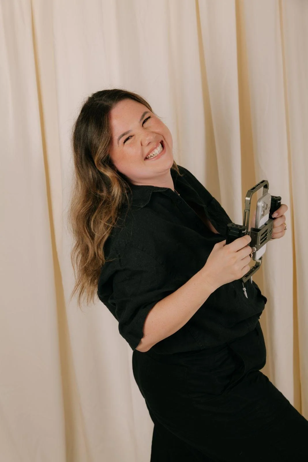 Femme souriante tenant un appareil photo, debout devant un rideau beige.