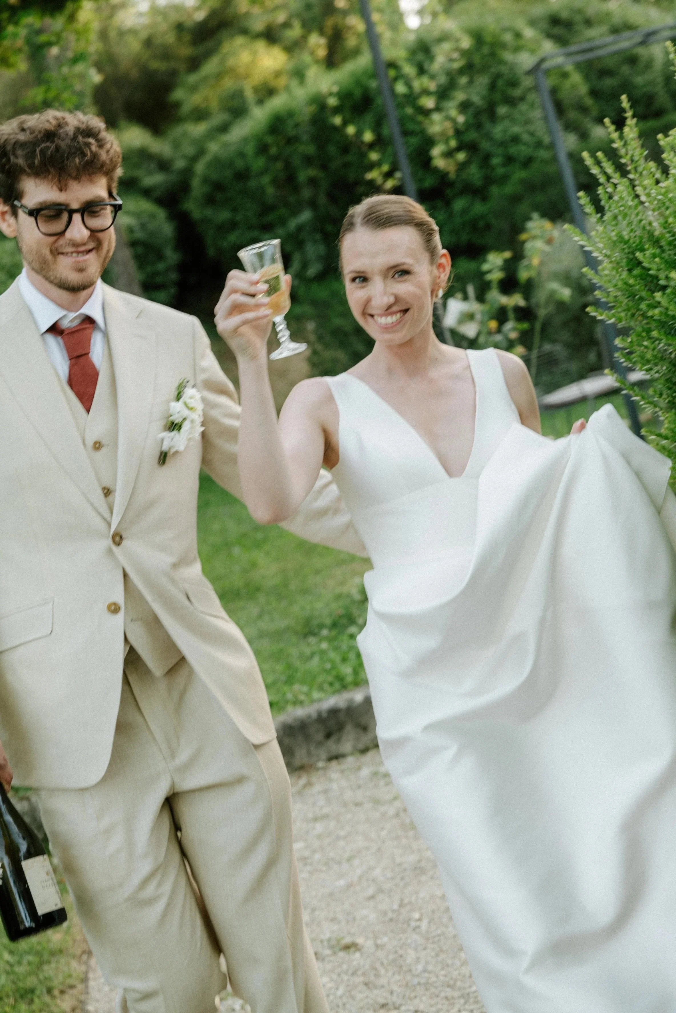 Un couple lors d'un mariage en plein air, la femme porte une robe de mariée blanche et tient un verre à vin, l'homme porte un costume clair avec une boutonnière blanche.