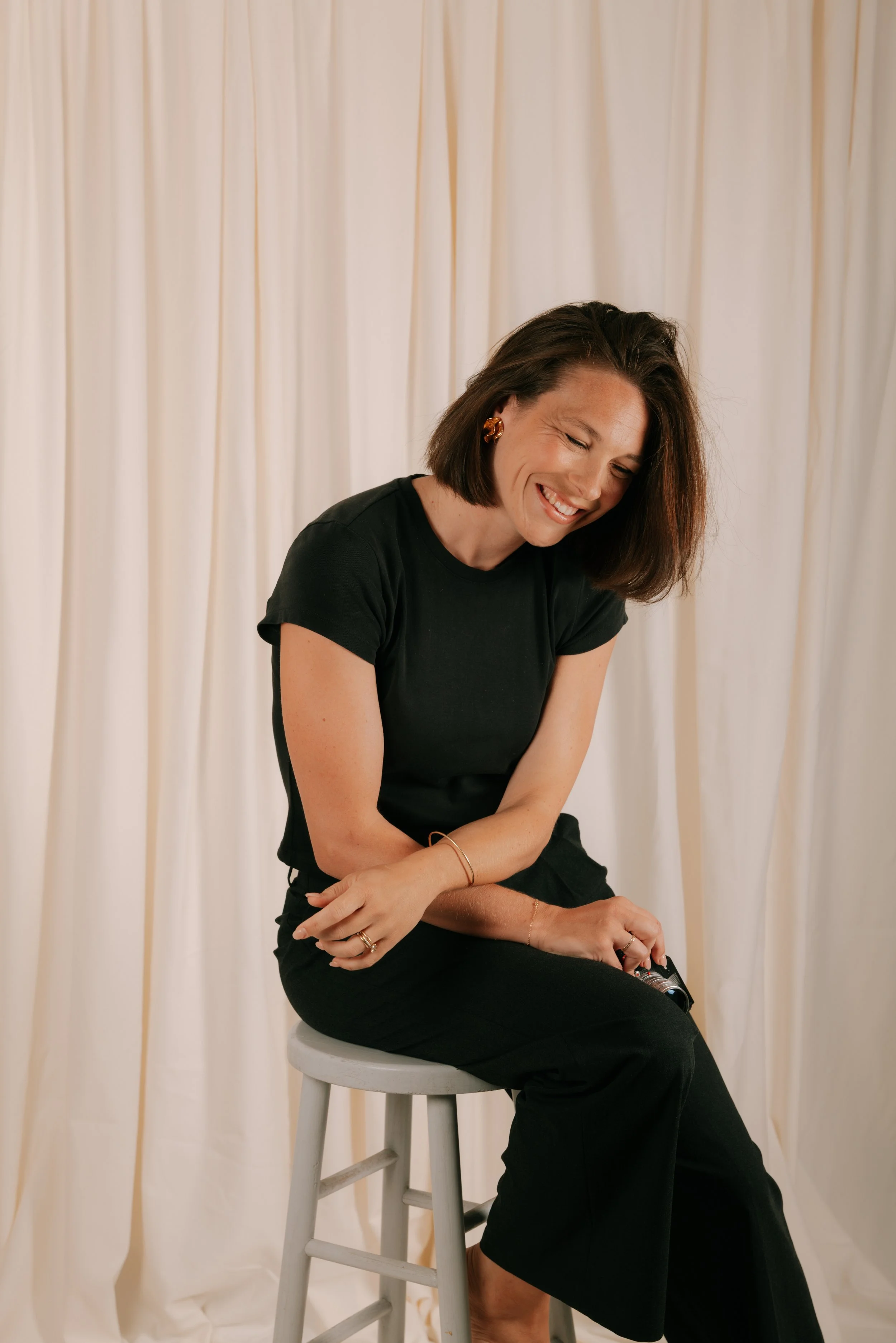 Femme souriante assise sur un tabouret blanc, portant un tee-shirt noir et des bijoux, avec des rideaux blancs en arrière-plan.