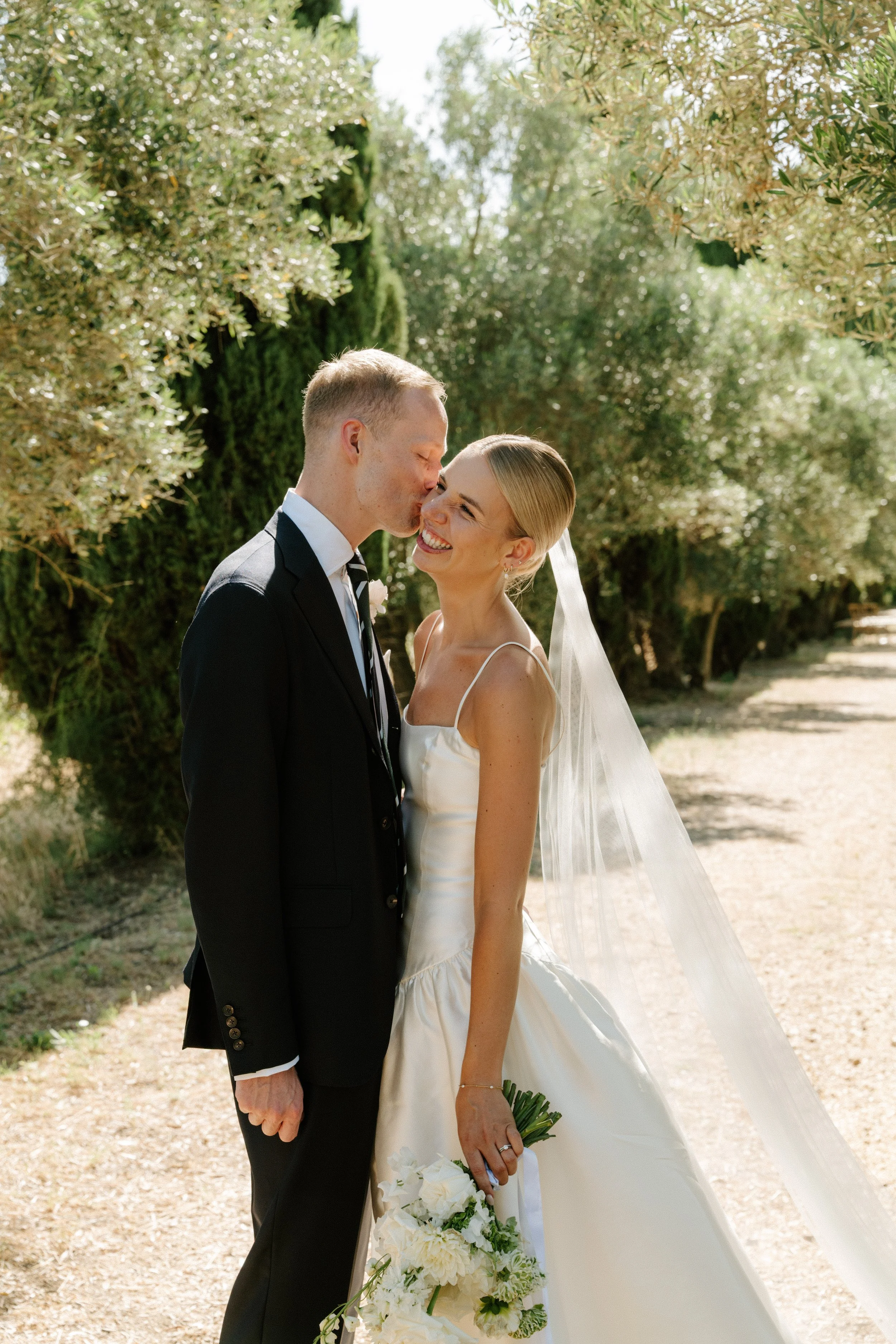 Un couple en tenue de mariage, la mariée tenant un bouquet de fleurs blanches, partage un moment tendre en souriant et en se touchant le front, dans un décor naturel avec des arbres et un chemin.