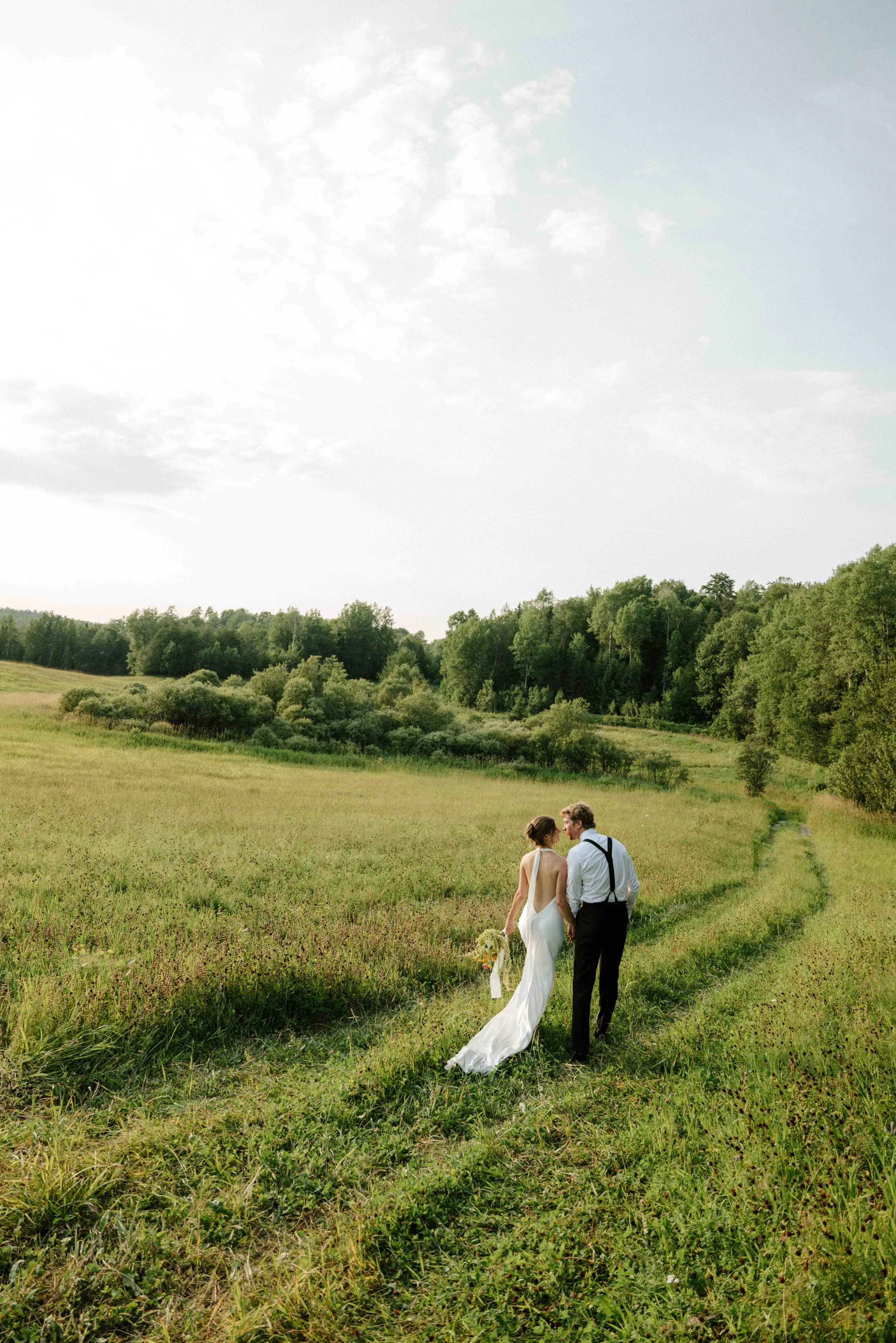 Un couple de mariage marche main dans la main dans un champ vert au milieu de la nature, avec un ciel partiellement nuageux au-dessus.
