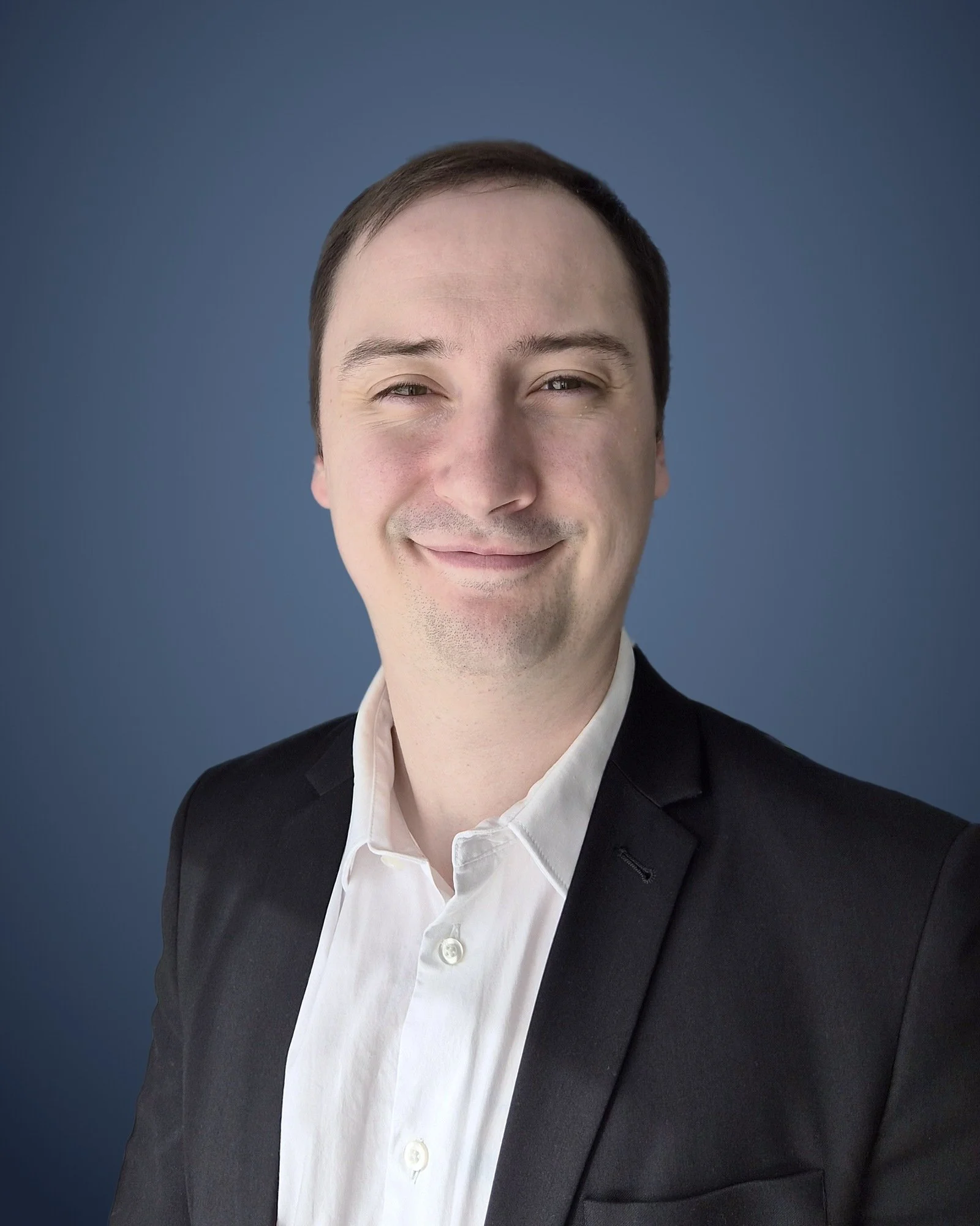 Professional headshot of a smiling man wearing a black blazer and white shirt against a blue background