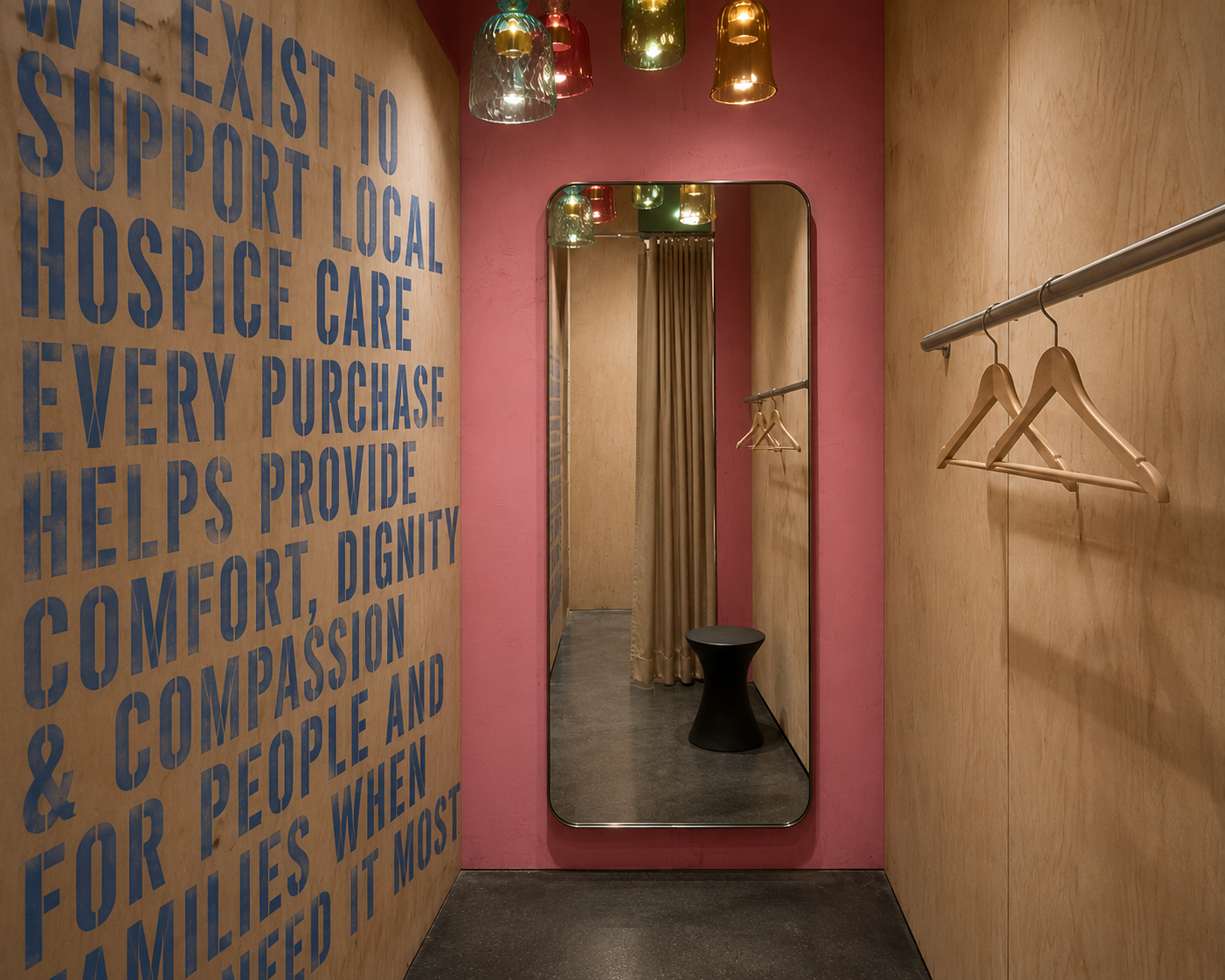 A small fitting room area with a full-length mirror, wooden walls, and a pink accent wall with large blue text. Wooden hangers hang on a metal rod, and colorful pendant lights hang from the ceiling. There is a black stool and beige curtains in the background.