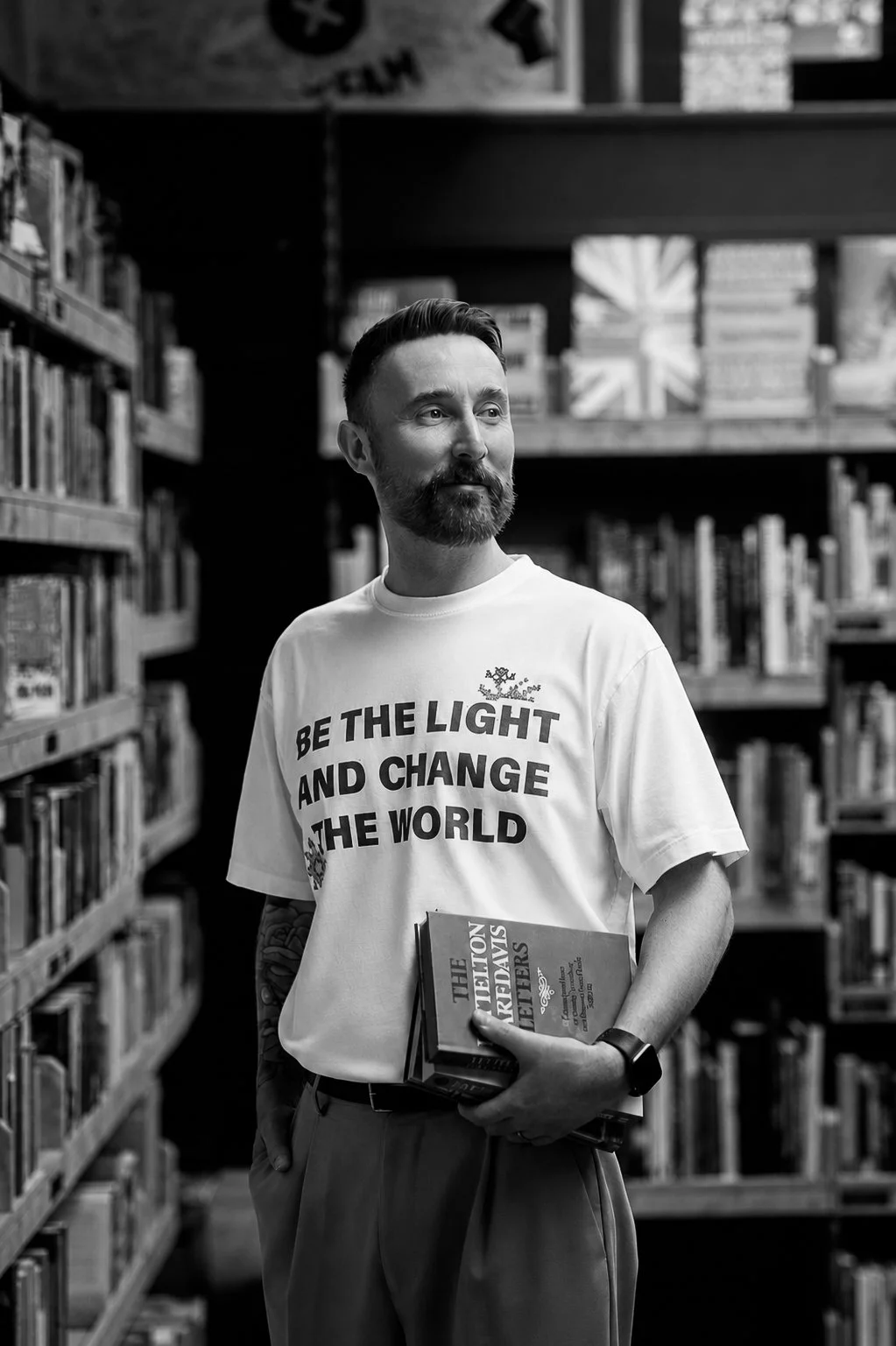 A man with a beard and mustache wearing a t-shirt that says 'Be the light and change the world' in a bookstore, holding a book titled 'The Melatonin Revolution.' The bookshelf behind him is filled with books, and a poster with a cross symbol is visible at the top.