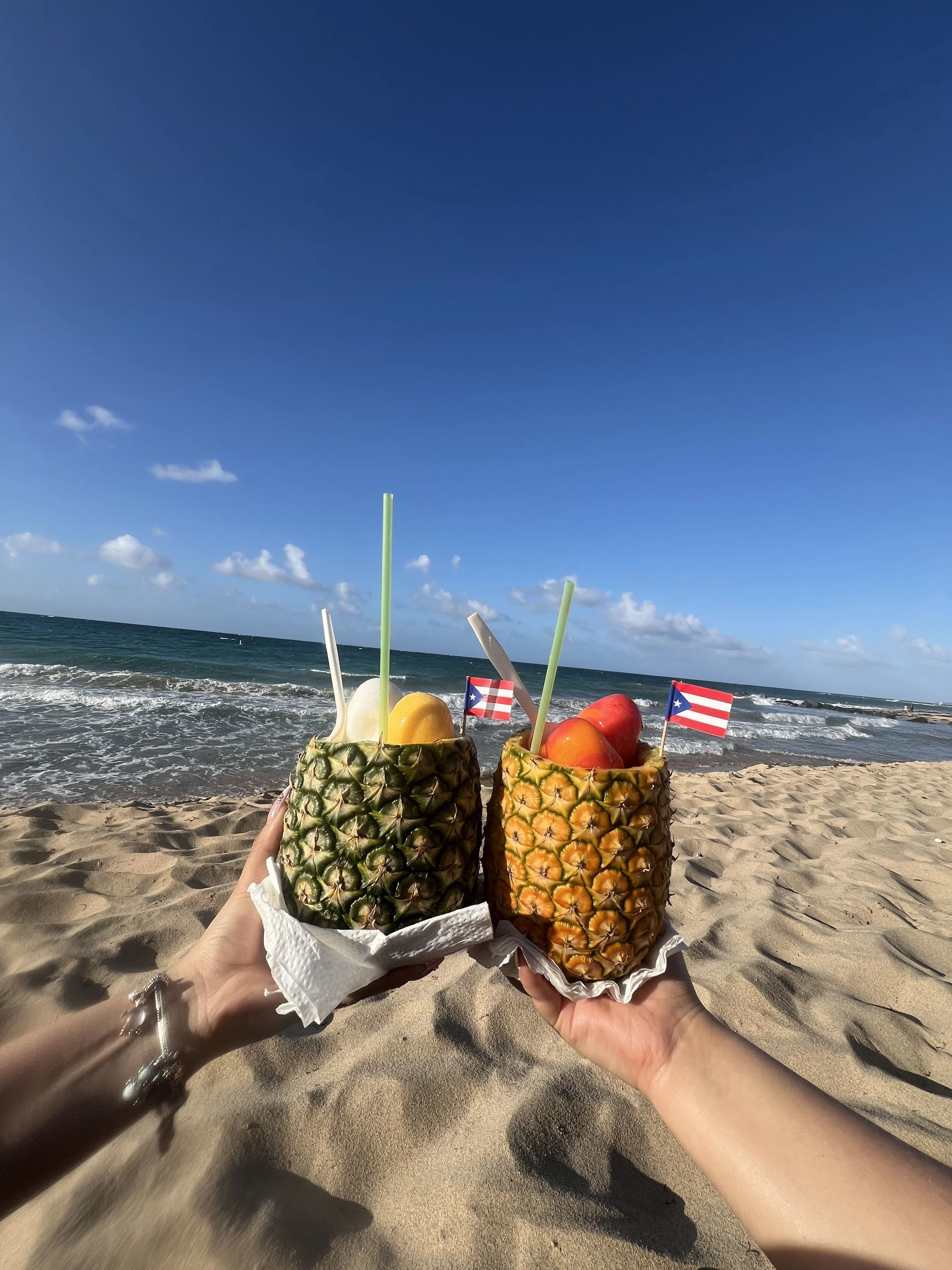 Two hands holding pineapple halves filled with fruit and drinks with straws and small Puerto Rican flags, on a sandy beach with the ocean and blue sky in the background.