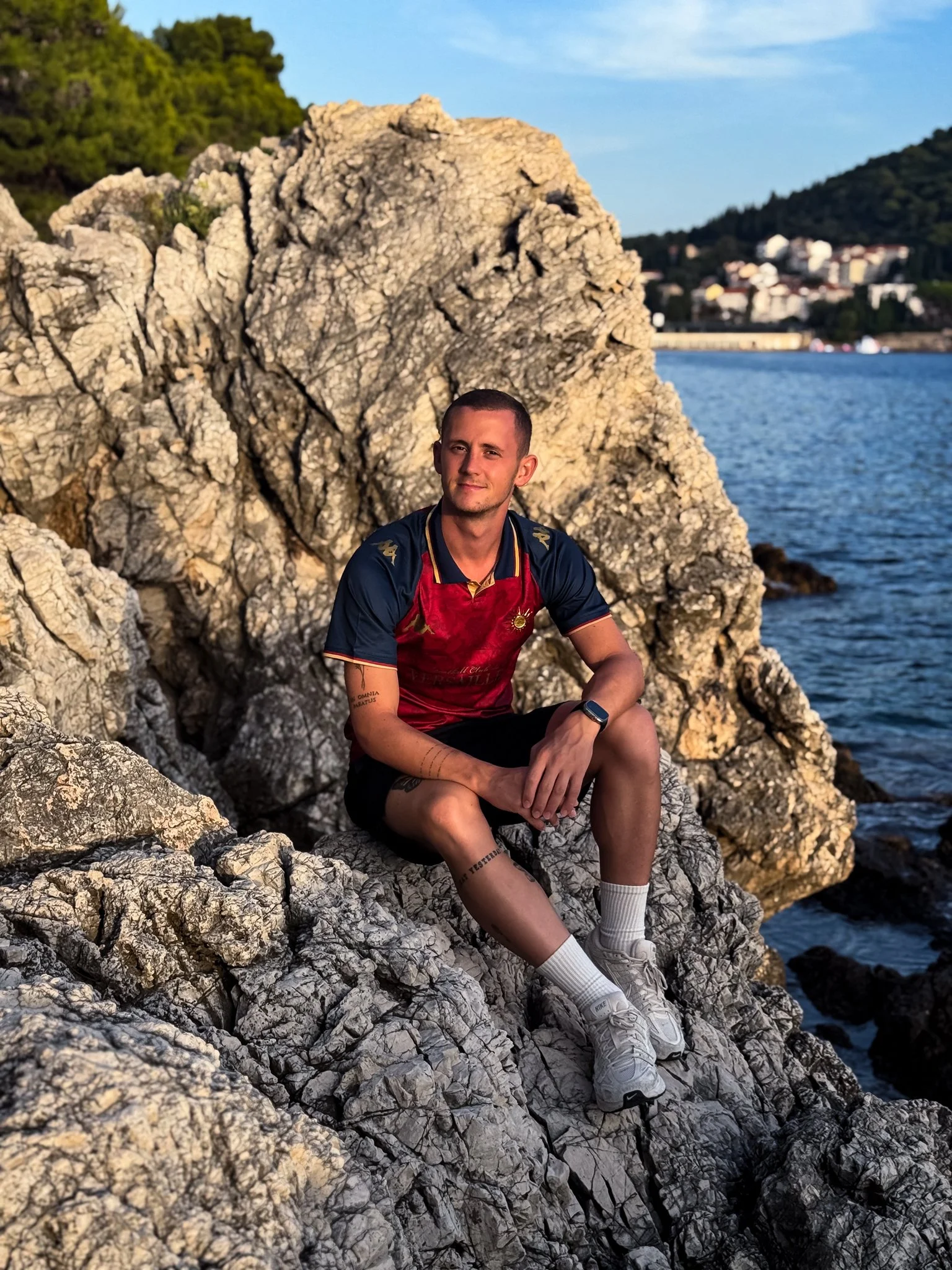 Un jeune homme assis sur des rochers au bord de l'eau, portant un pantalon court, des baskets blanches, un t-shirt sportif et une montre. En arrière-plan, il y a une mer, une colline avec des bâtiments, et le ciel bleu.