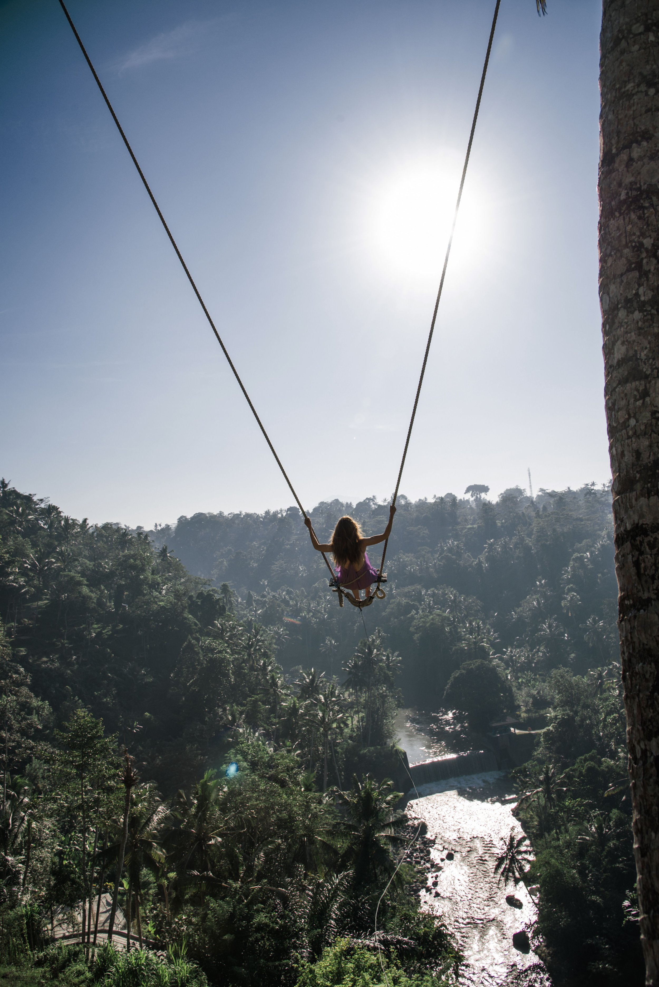 A girl on a swing high above a river and lush forest, with the sun shining brightly in the clear sky.