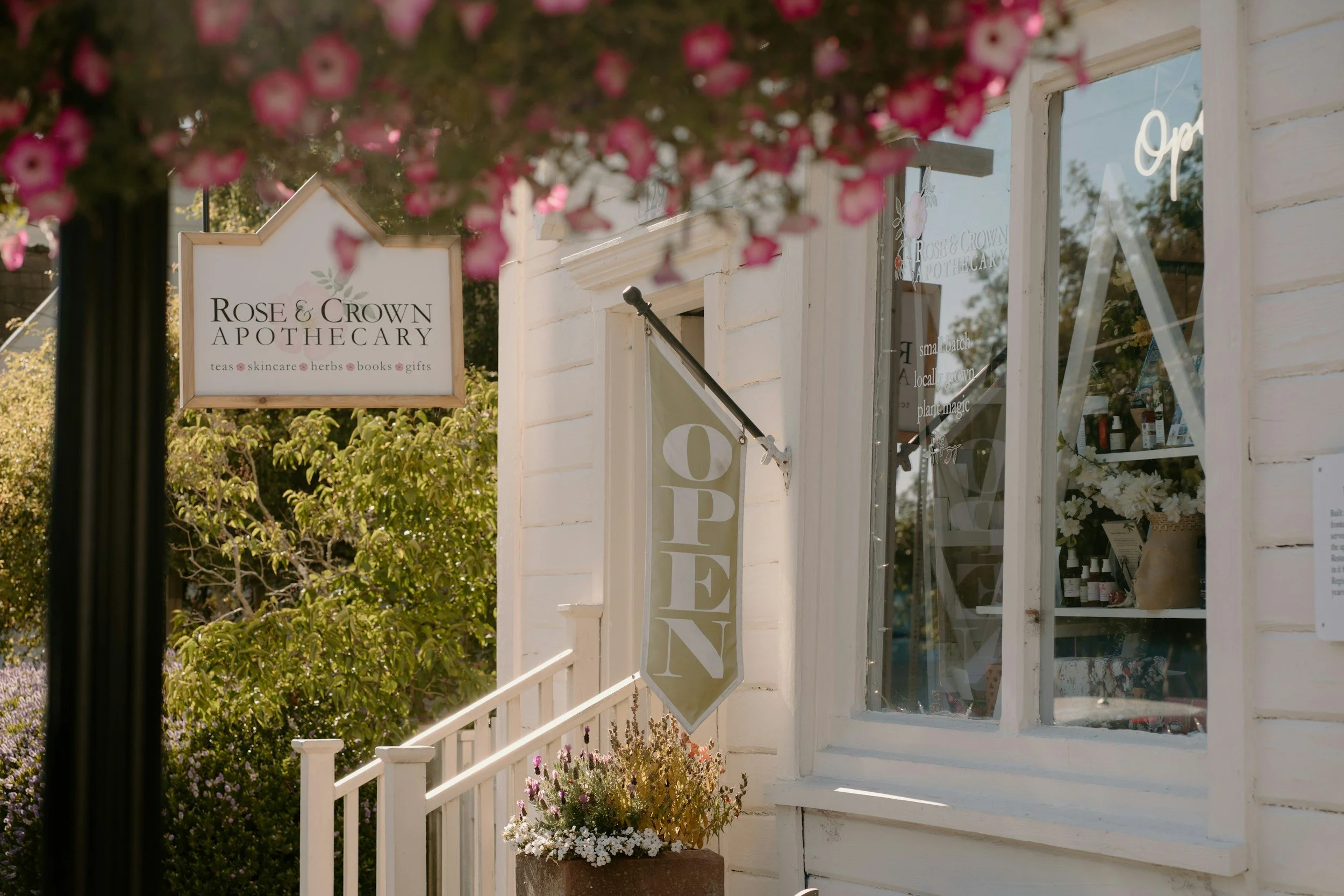 Exterior of a shop called Rose & Crown Apothecary with a white sign, flowers in a pot, and window displaying products, surrounded by pink flowers in the foreground.