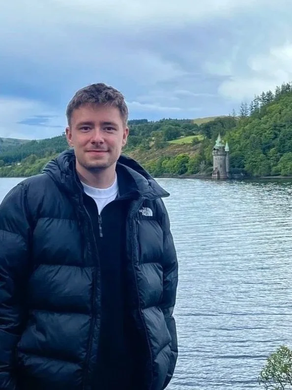 Young man standing outdoors near a body of water, wearing a black puffy jacket, with greenery, hills, and a castle-like structure in the background under a cloudy sky.