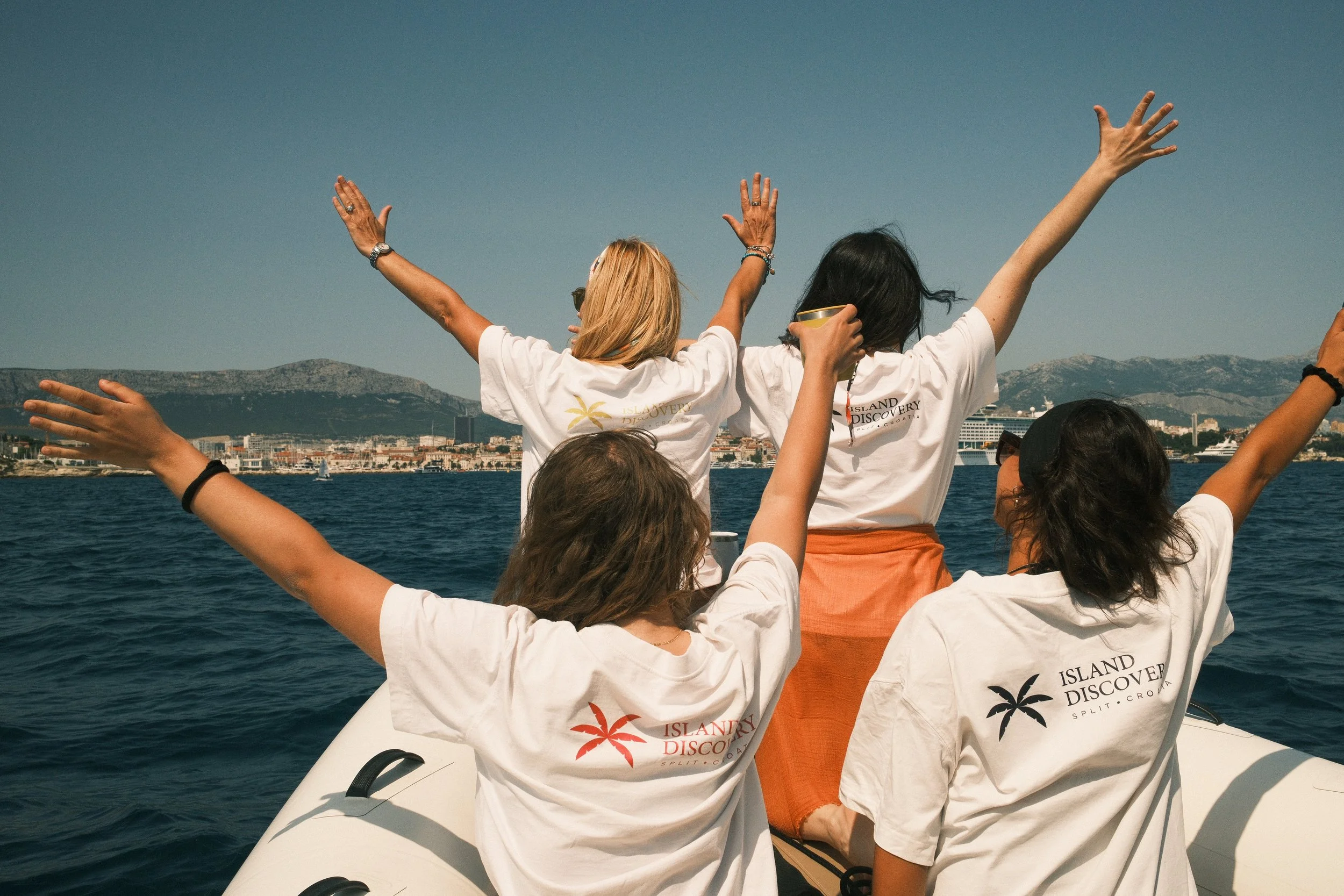 Group of women on a boat with arms raised, wearing white T-shirts that say 'Island Discovery,' with water and mountains in the background.