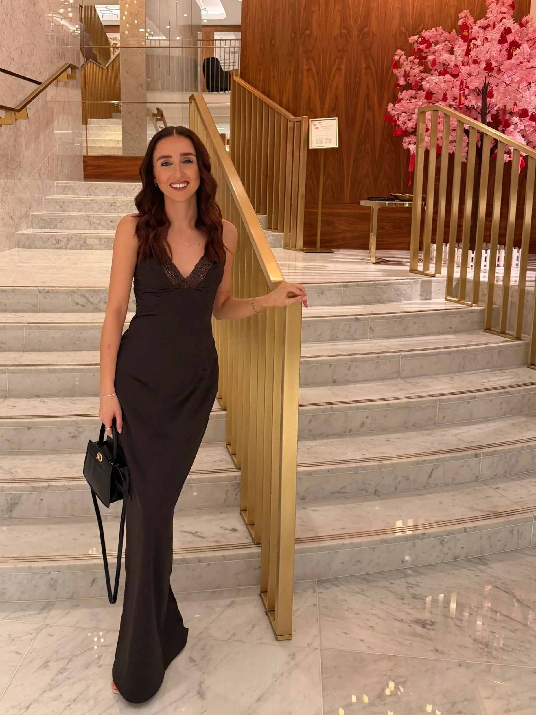 A woman dressed in a black dress standing on a marble staircase inside a building, smiling at the camera. She is holding a black handbag and has wavy brown hair. Behind her, there are wooden railings, a pink artificial cherry blossom tree, and a small table.