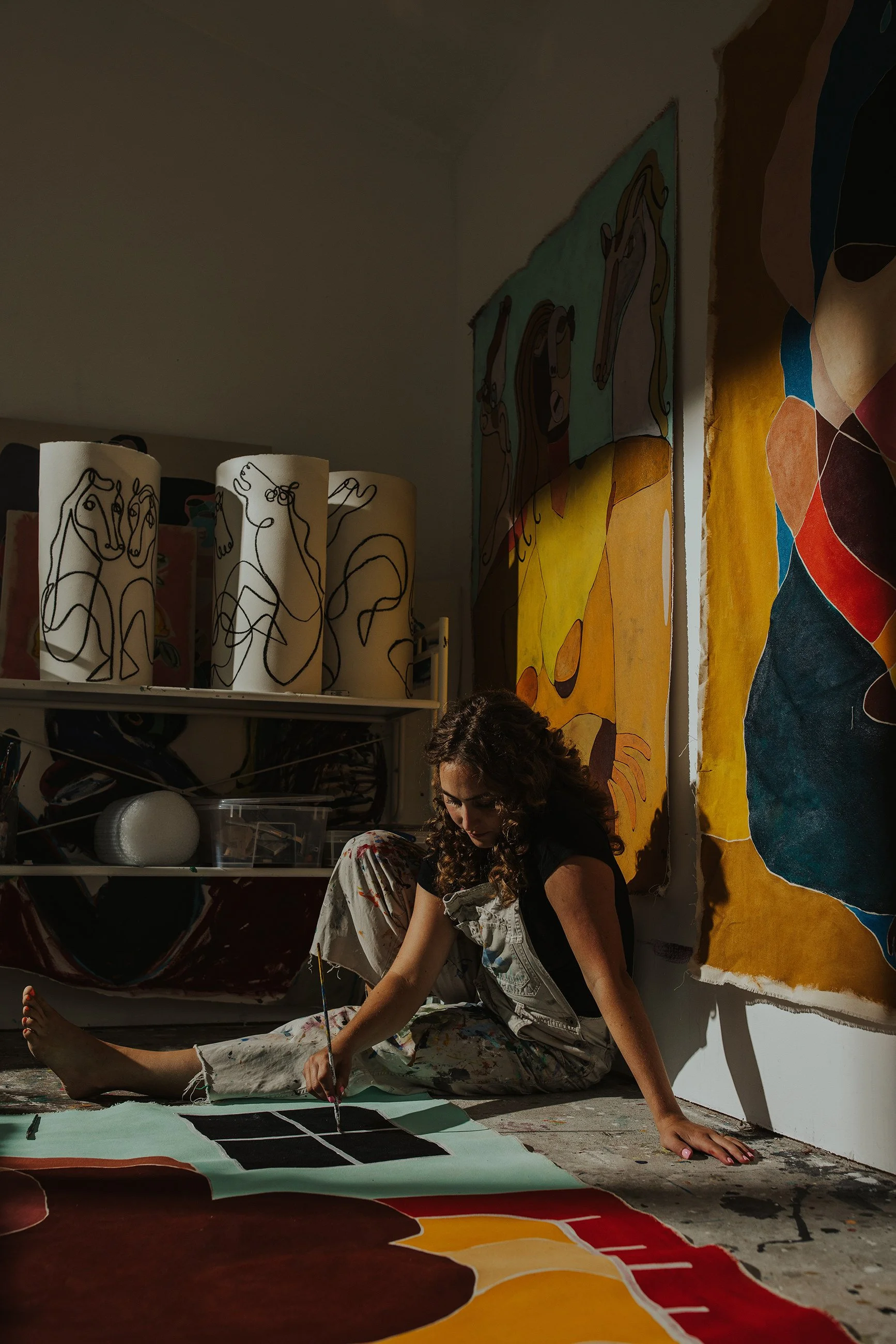 A woman with curly hair wearing paint-stained overalls sitting on the floor in an art studio, working on a large canvas with abstract colorful designs and paintings on the wall behind her.
