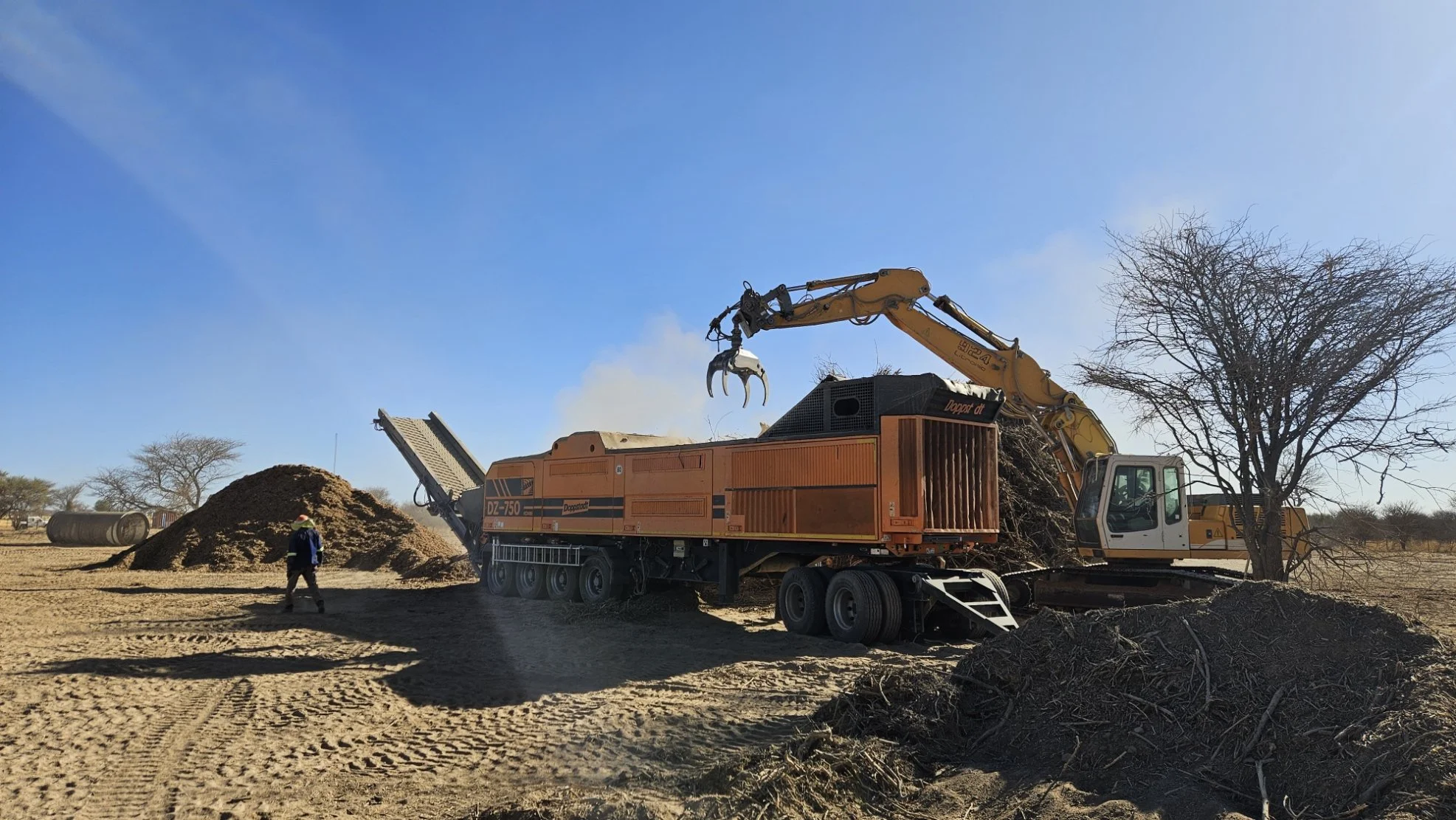 An excavator loading dirt into a mobile crusher to produce gravel on a construction site; a worker in a blue safety vest and helmet observes nearby. Trees and a blue sky are visible in the background.