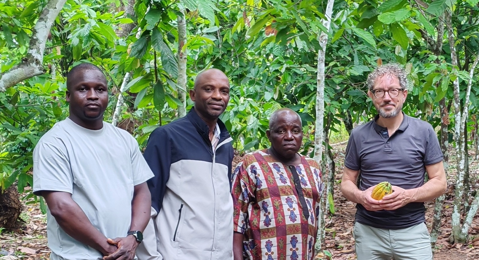 Four men standing in a lush green forest, with one man holding a yellow and green cacao pod.