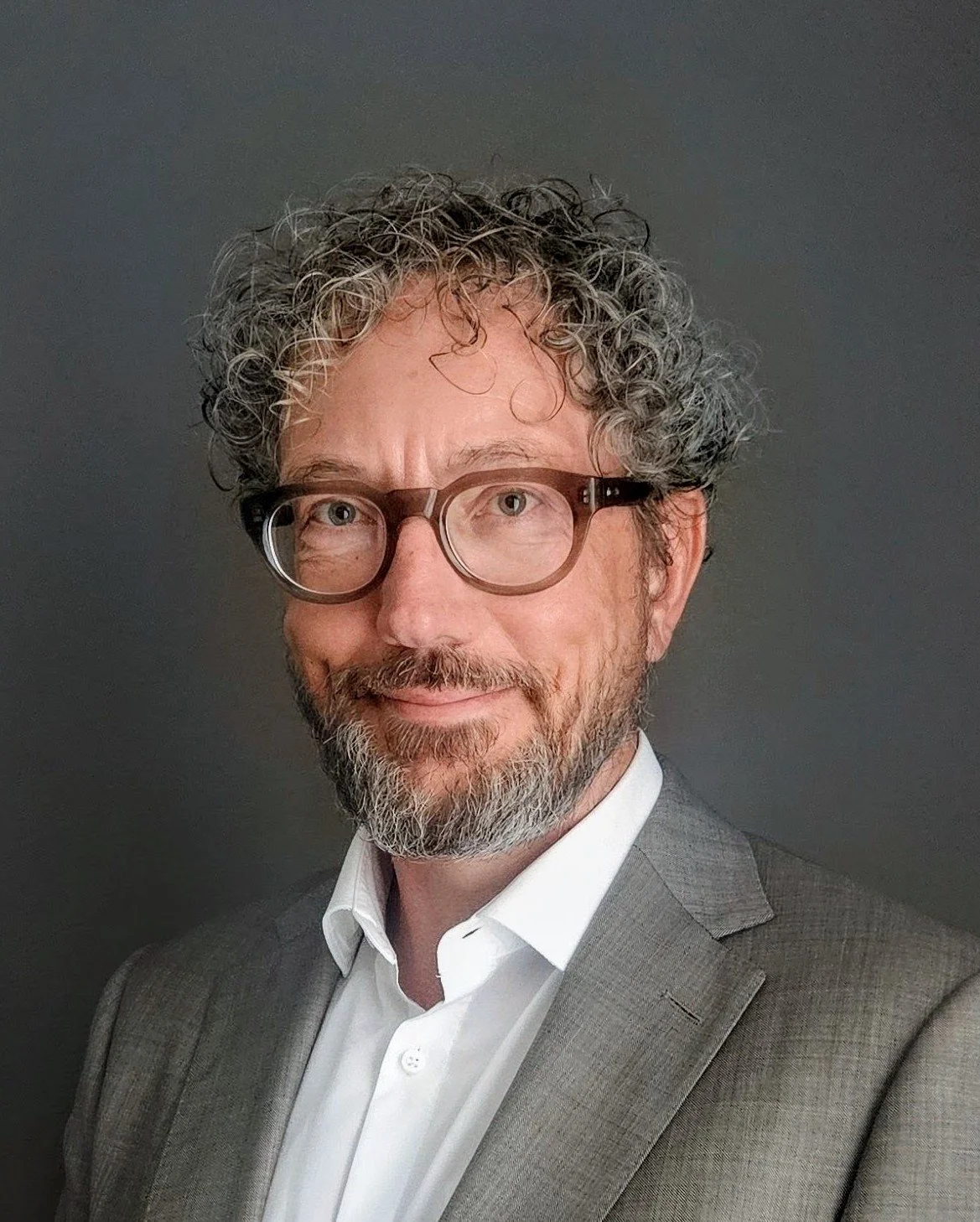 A man with curly gray hair, glasses, and a beard wearing a gray suit and white shirt.