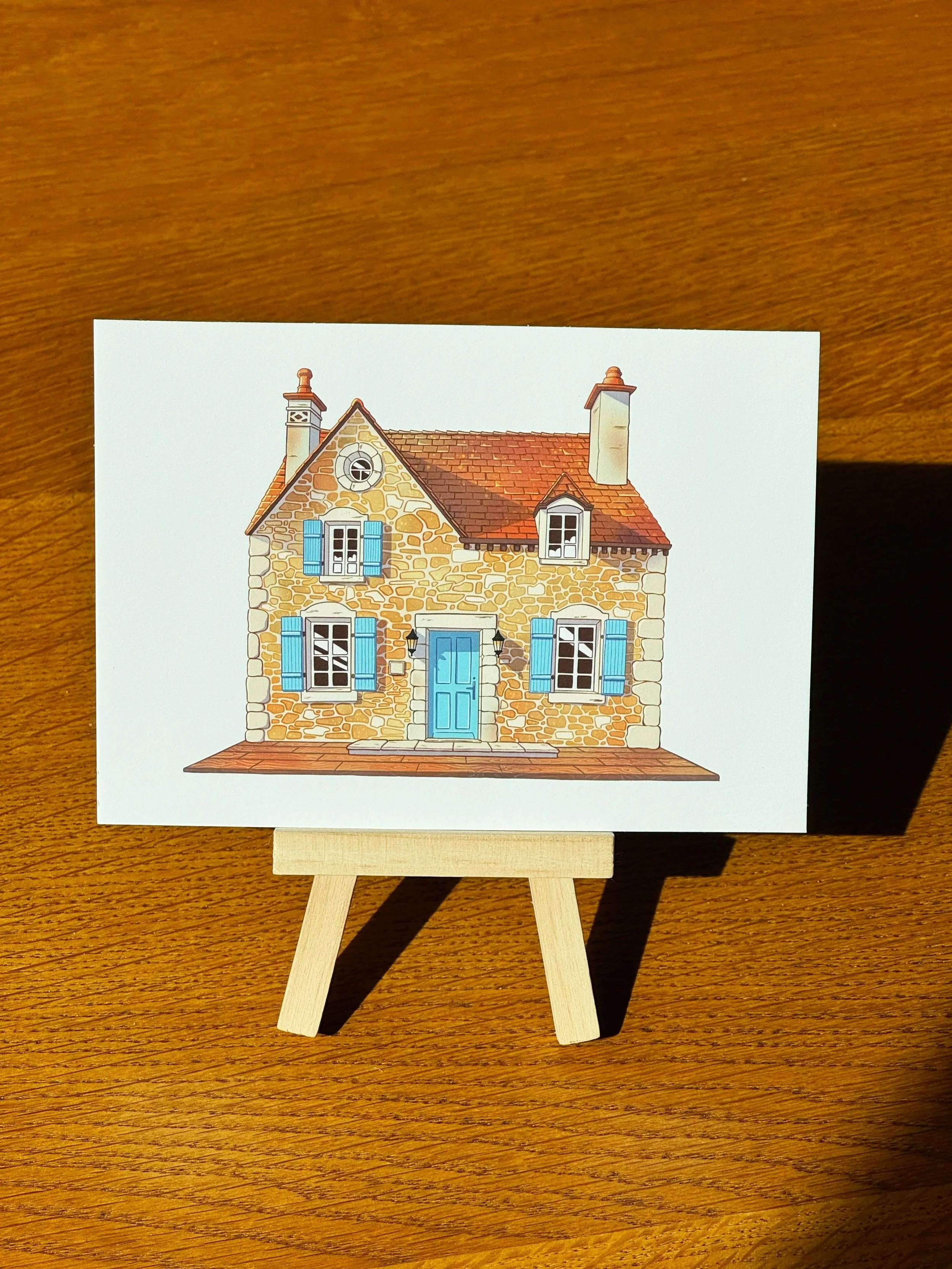A print of a house with stone walls, a red-tiled roof, blue shutters, and a blue door on a small easel on a wooden surface.