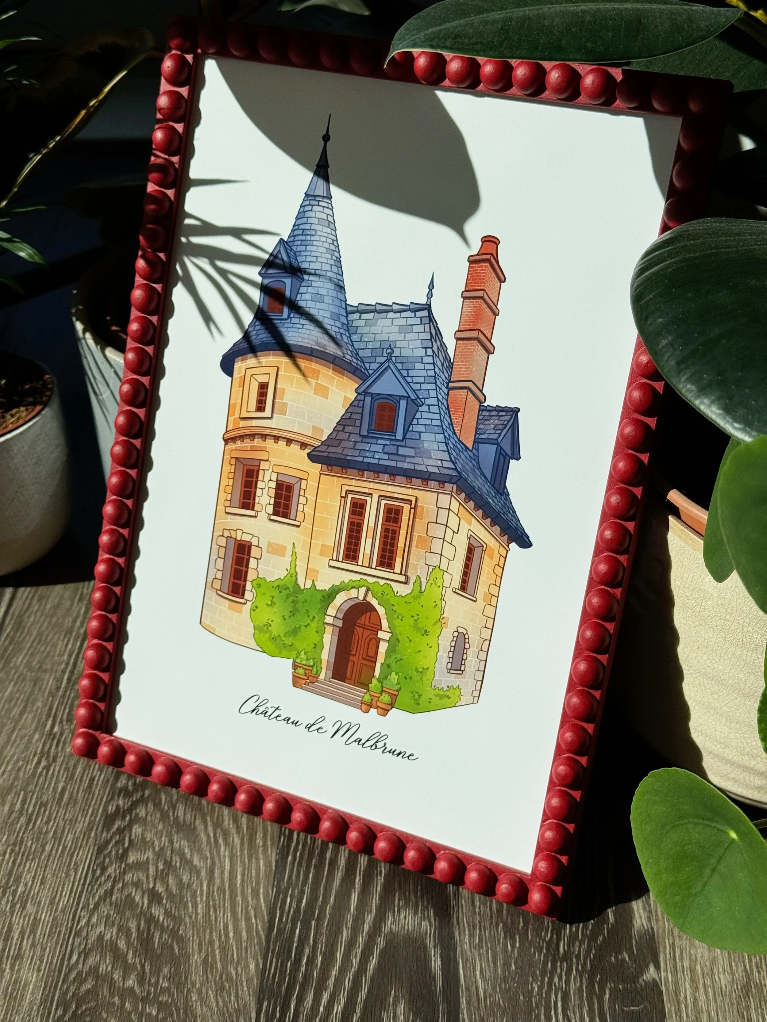 A framed illustration of a whimsical castle with pointed blue roofs and a tall brick chimney, surrounded by green shrubbery. The frame is decorated with red beads and the text below the illustration reads "Château de Malbrune."