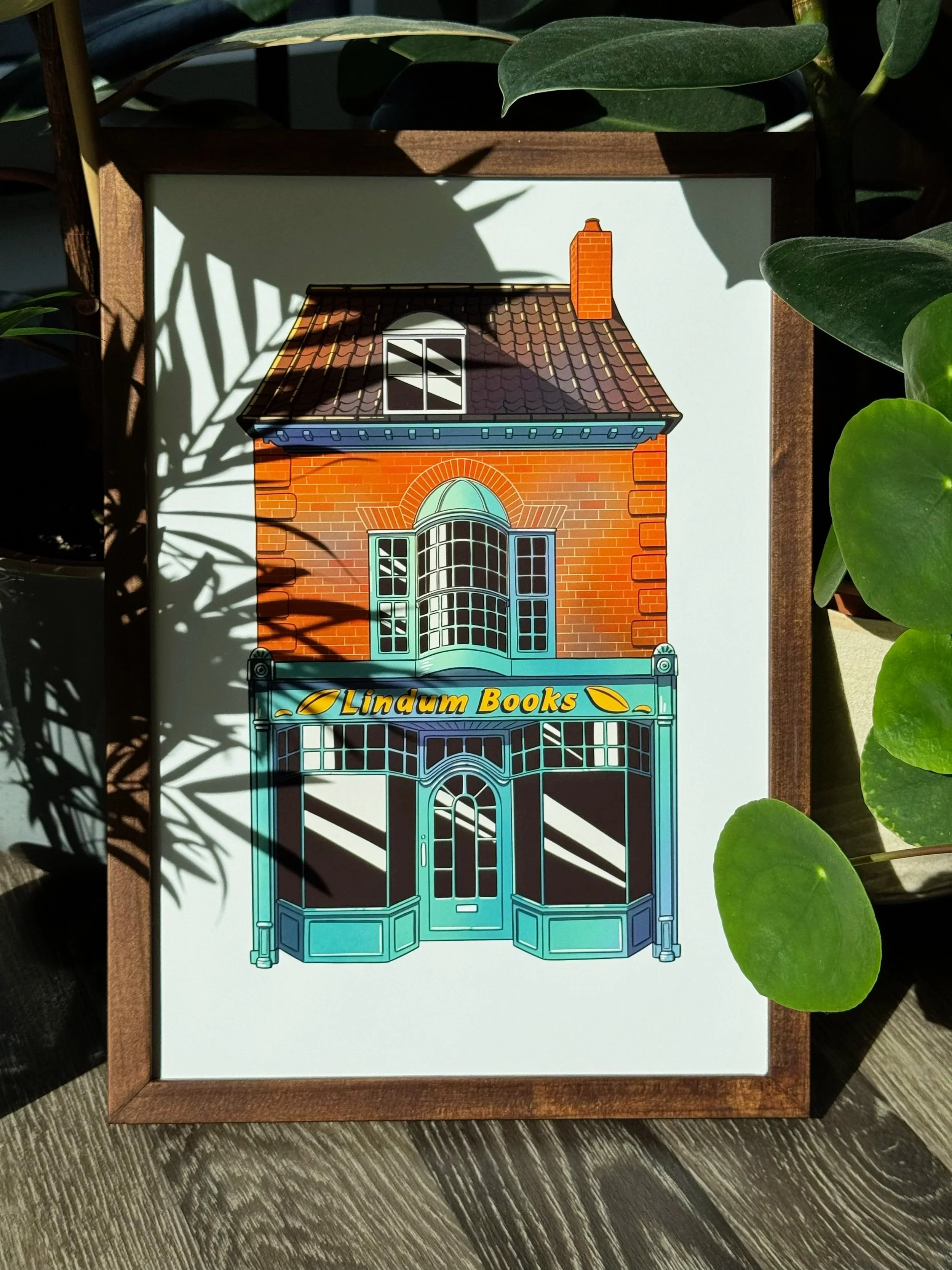 Framed illustration of a brick building facade with a large rounded window and blue storefront, with yellow text reading "Lindum Books" and decorative leaves.