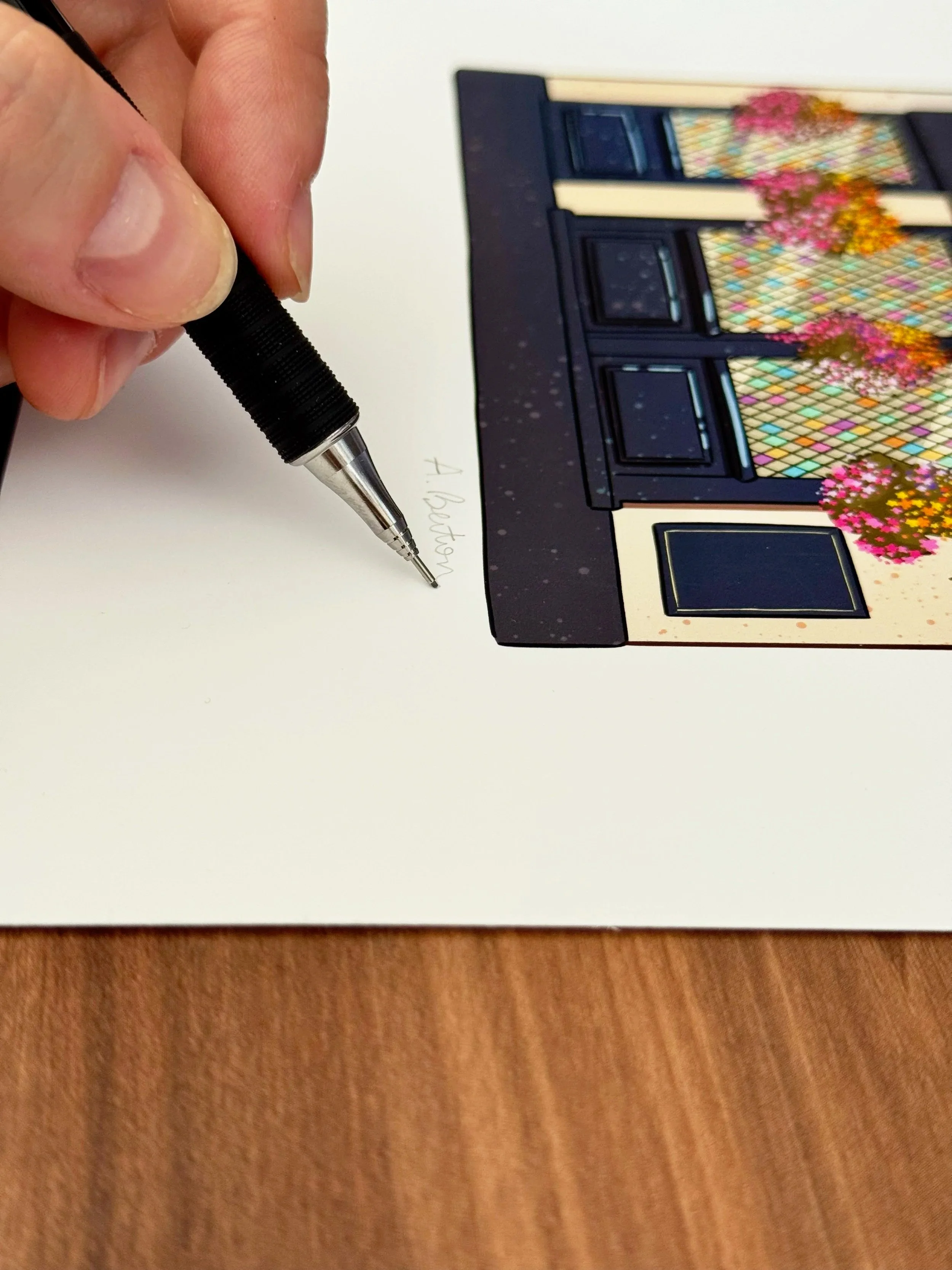 Close-up of a hand holding a black pen, writing on a white piece of paper. Part of a colorful illustrated building with flowers is visible in the background.