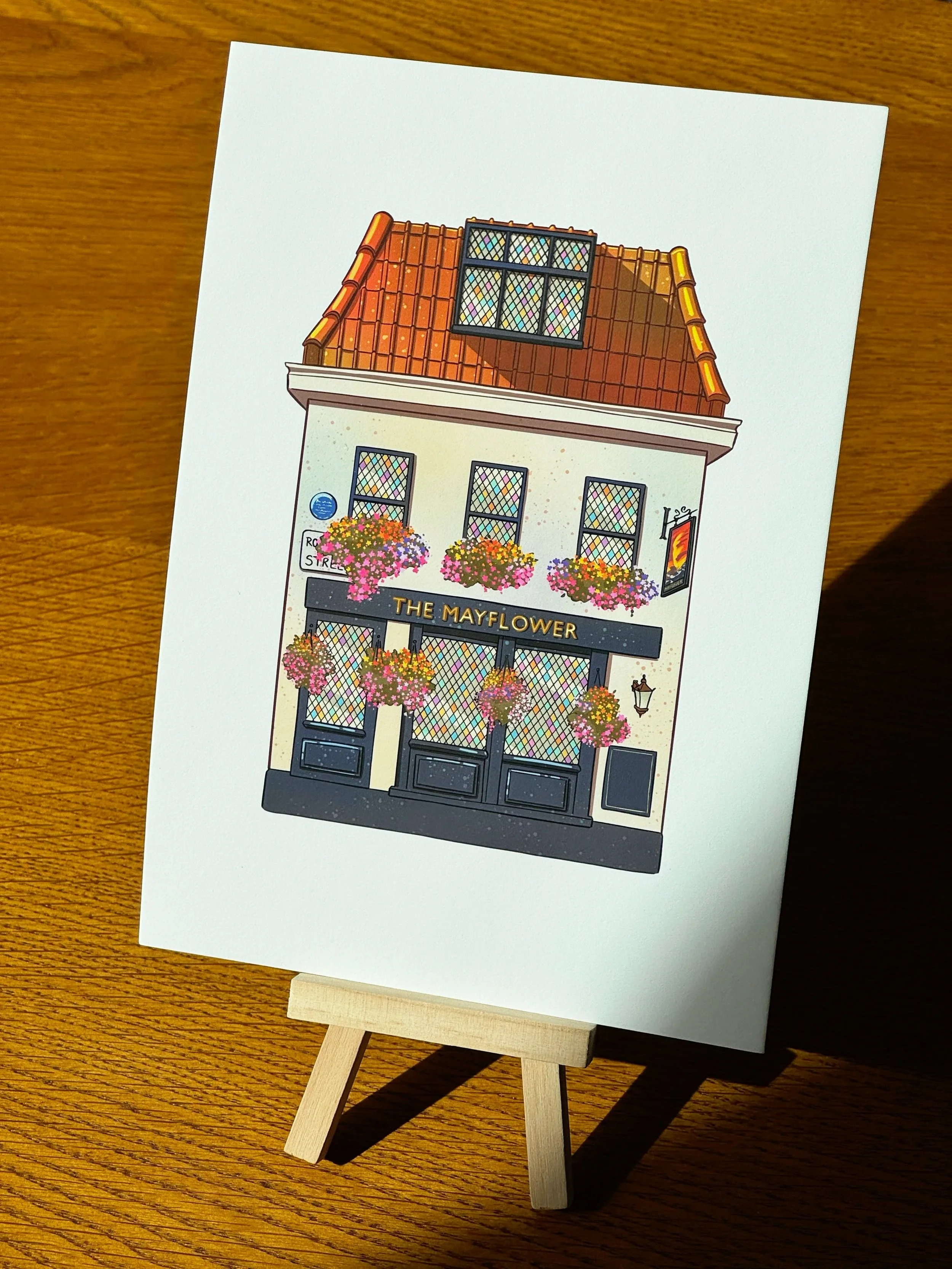 Illustration of a pub with a sign that reads 'The Mayflower', decorated with hanging flower baskets and a large window on the upper floor, set on a wooden table.