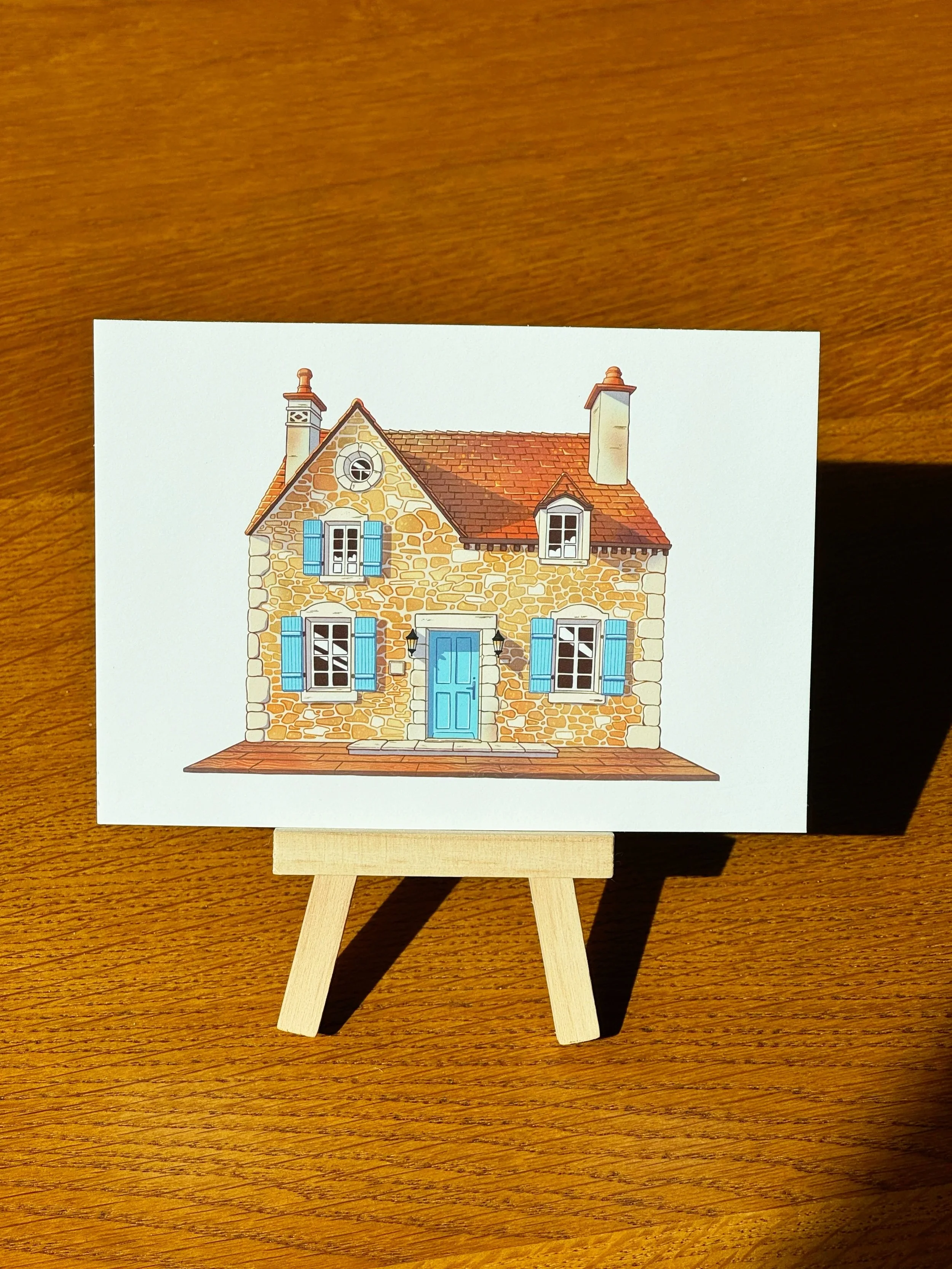Illustration of a stone house with blue shutters, a blue door, and a small front porch, displayed on a small wooden easel on a wooden surface.