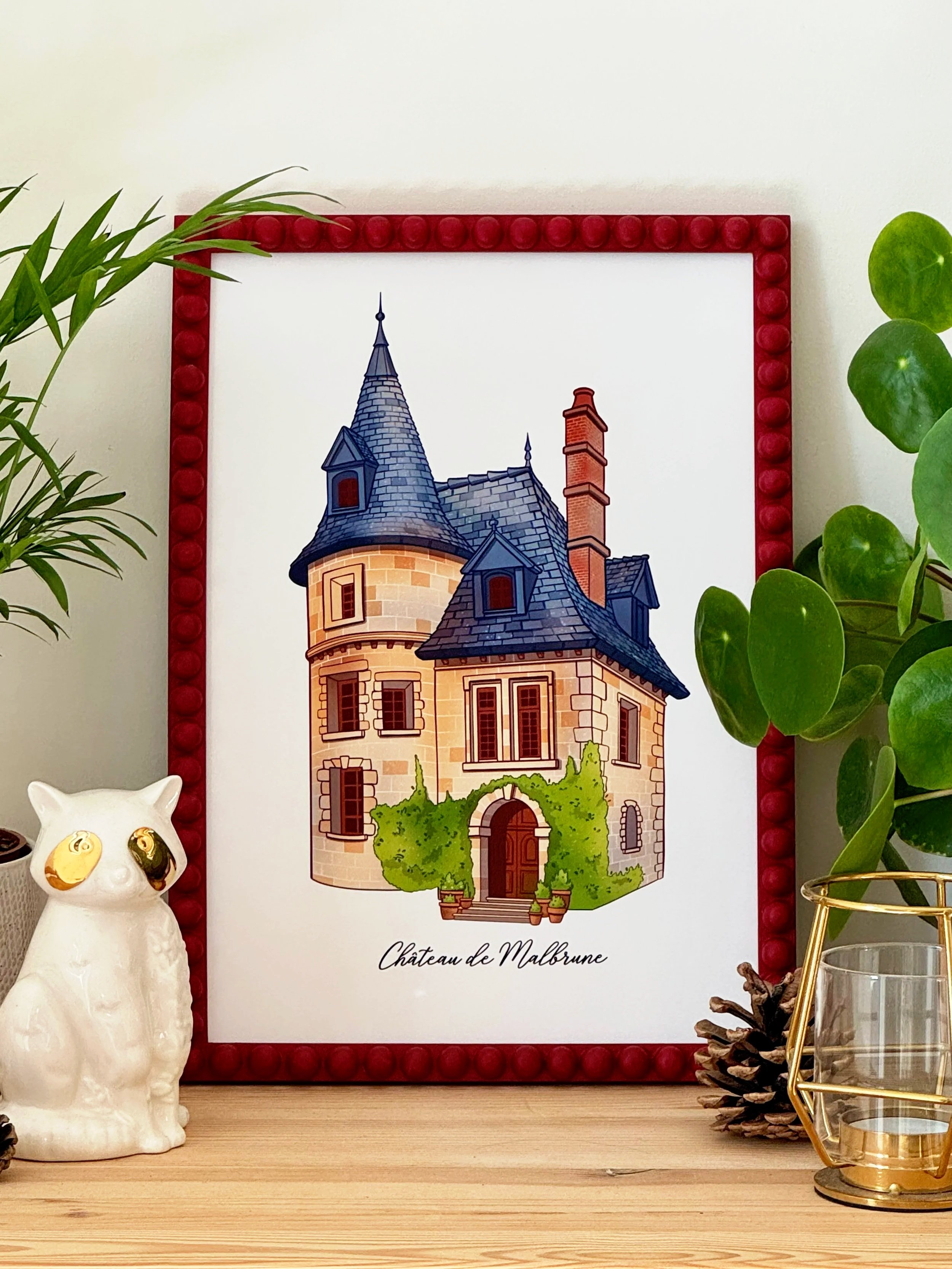 Decorative framed print of a castle labeled Château de Malbrune, surrounded by plants, pine cone, and candle holder with pine cone decoration on a wooden surface.