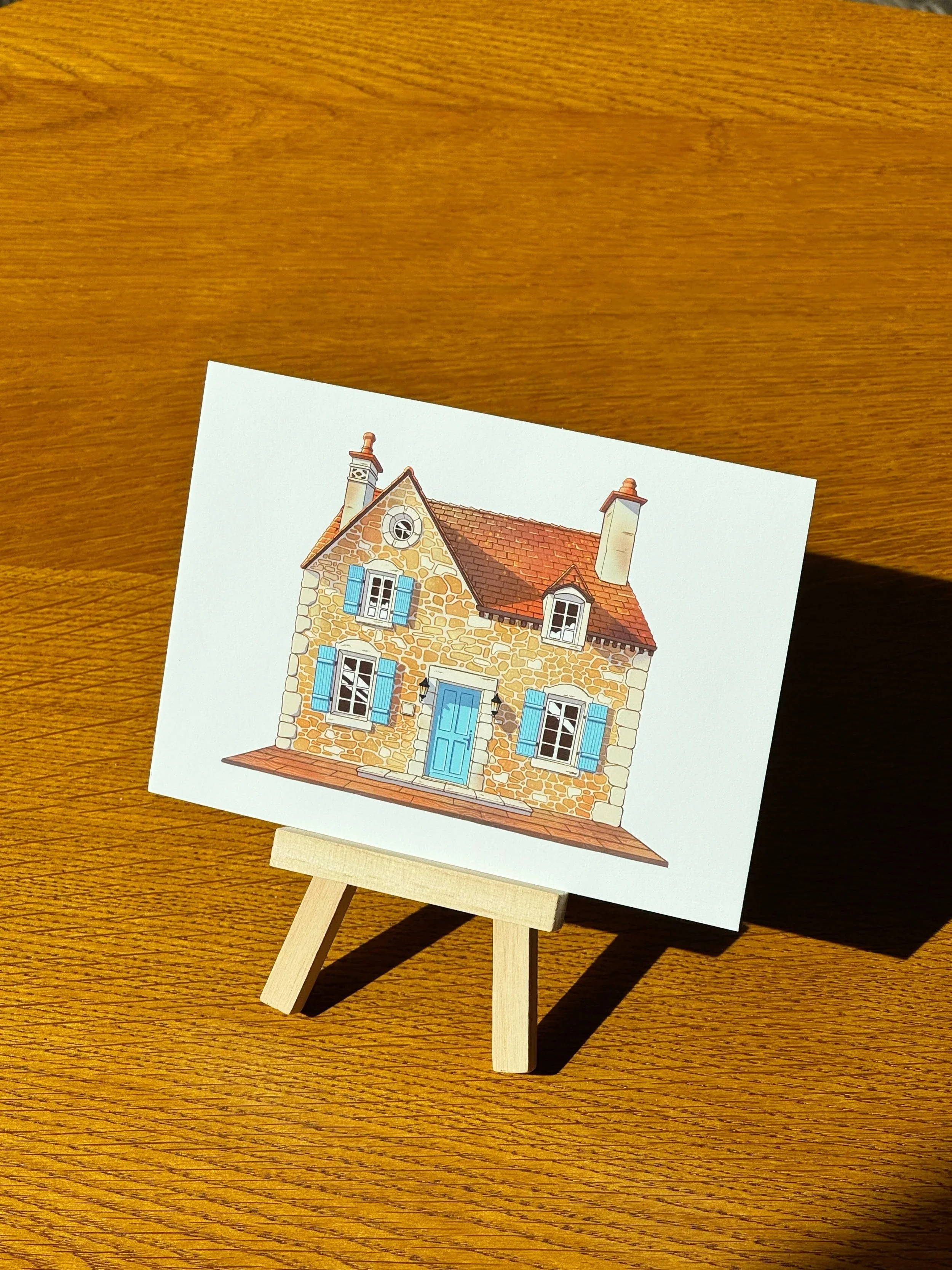 A printed illustration of a house with stone walls, blue shutters, and a red roof, displayed on a small easel on a wooden surface.
