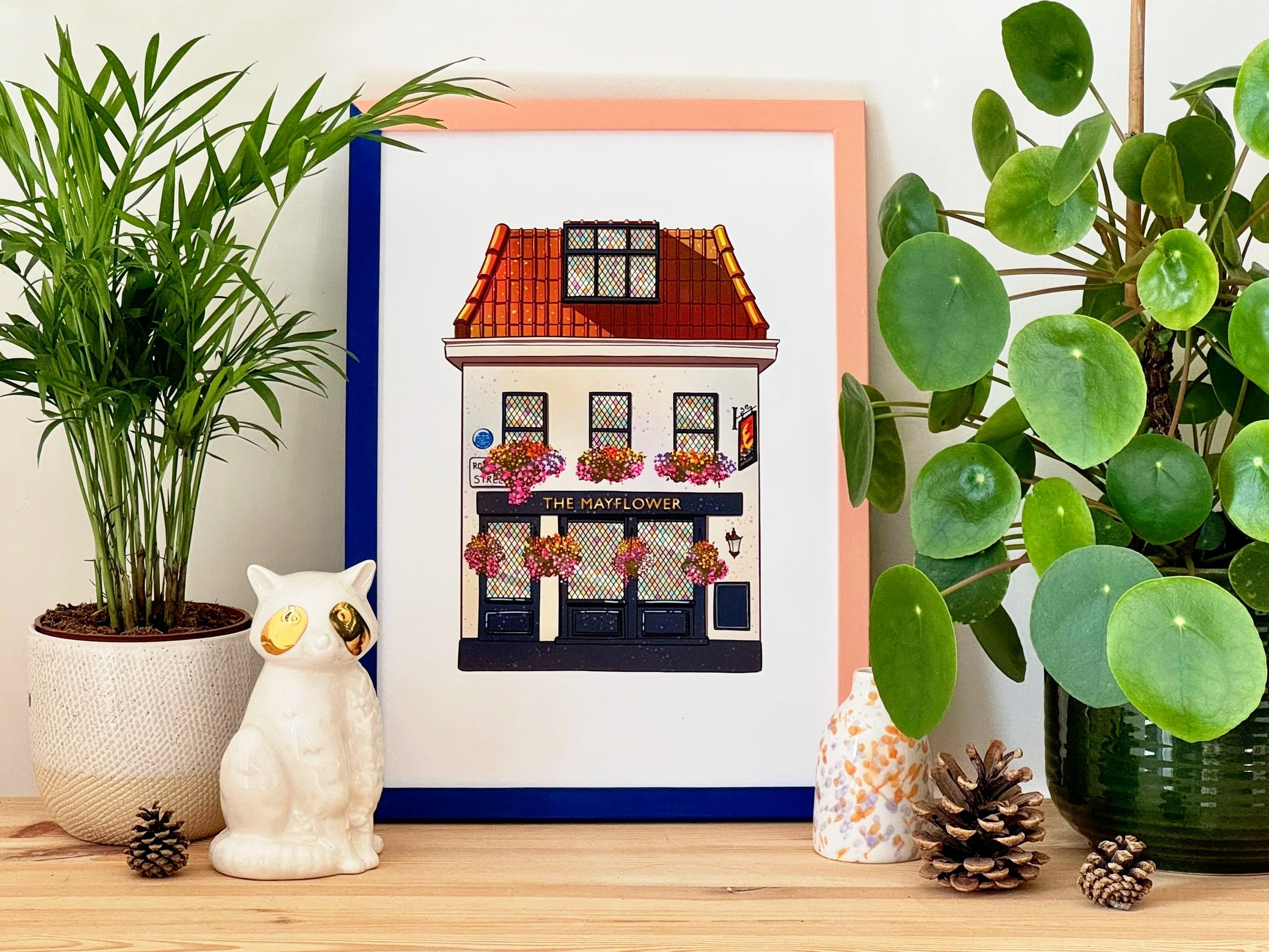Decorative display of houseplants, pinecones, and ceramic animal figurines on a wooden surface, with a framed illustration of a building labeled 'The Mayflower' in the background.