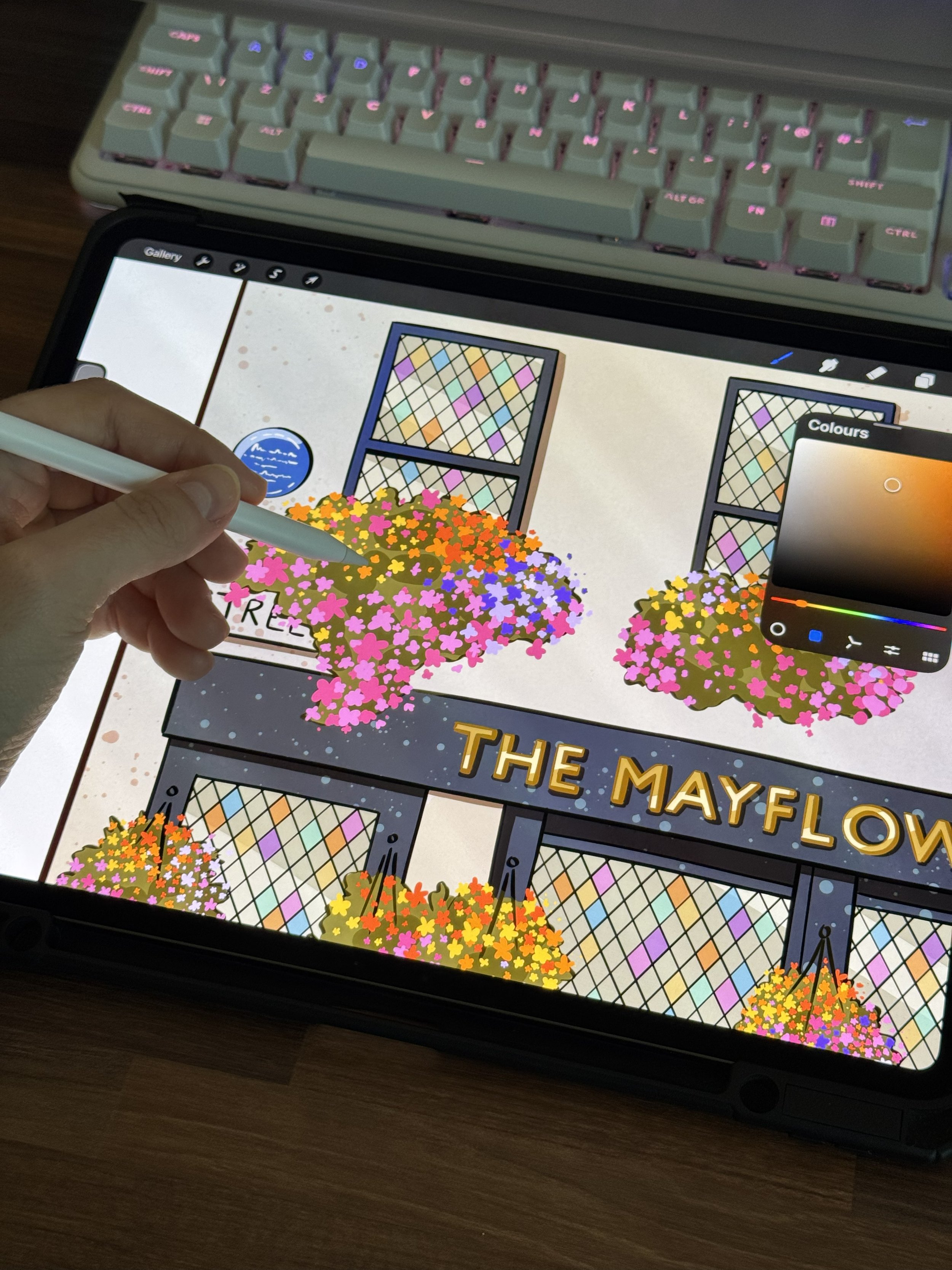 Close-up of a tablet screen displaying colorful flower illustrations, with a person holding a white stylus. A pink and white mechanical keyboard with backlit keys is also visible in the background.