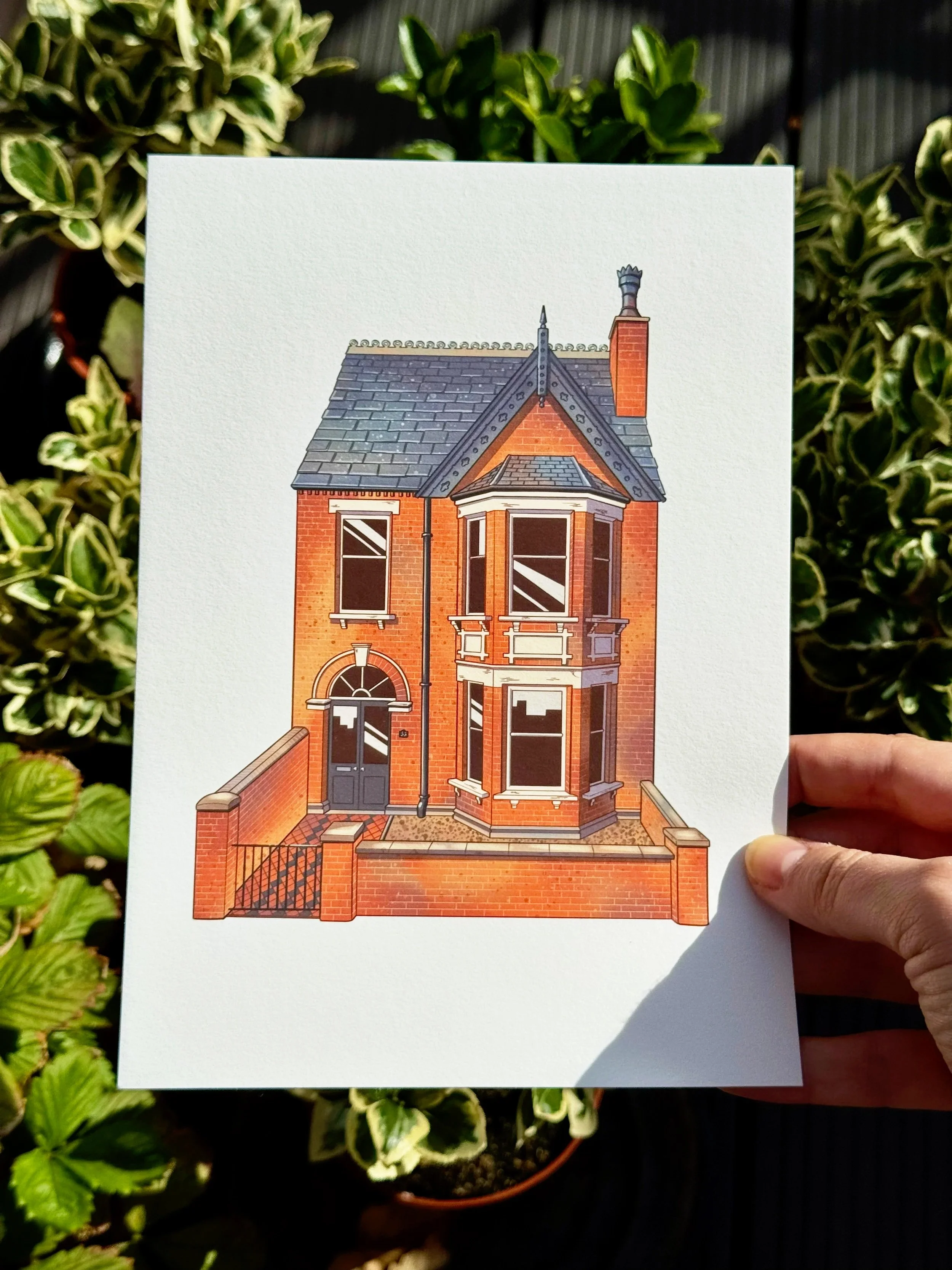 Illustration of a brick Victorian-style house with a bay window, arched front door, and a decorative roof, held in a person's hand outdoors with green plants in the background.