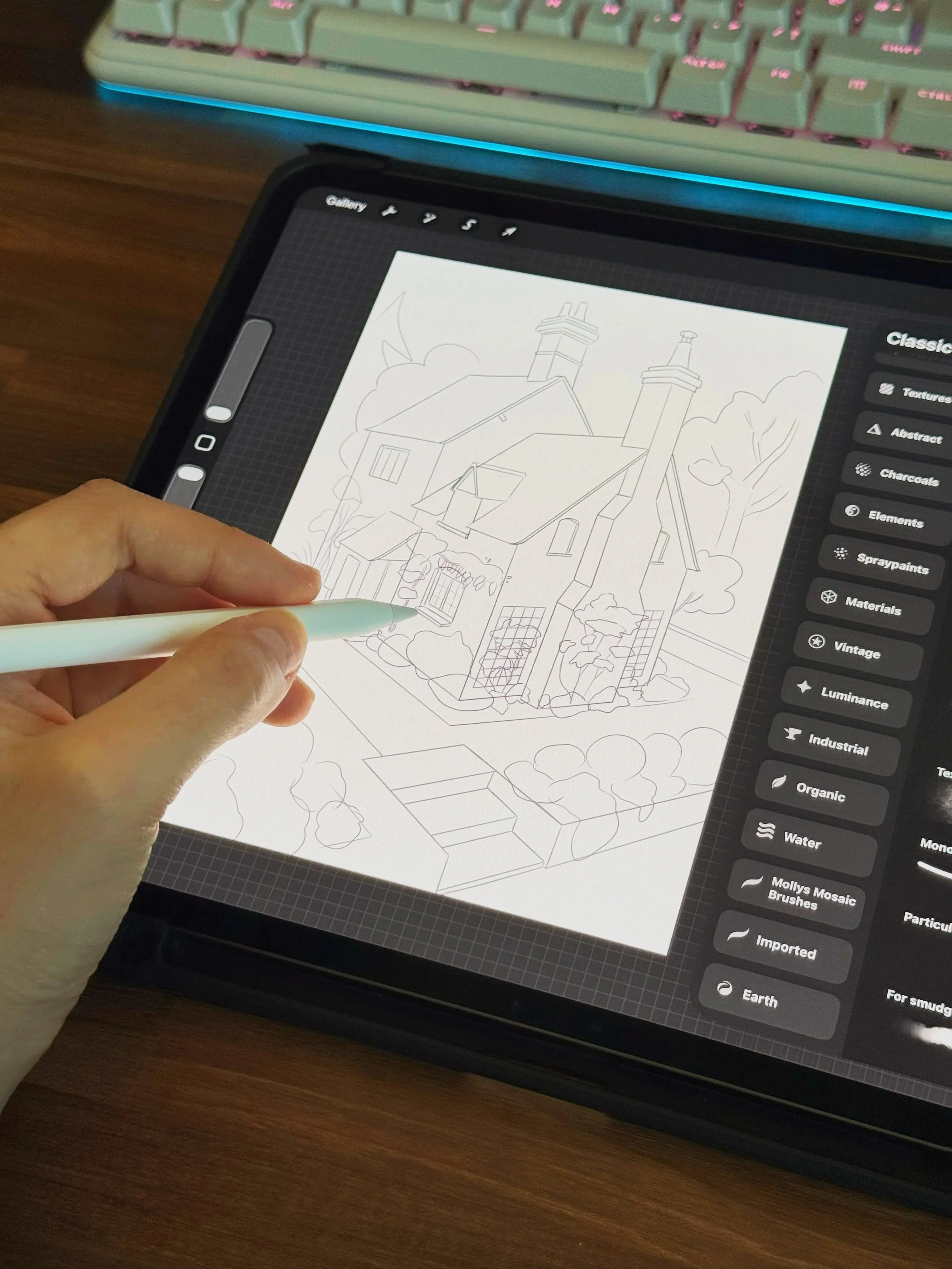 Digital tablet displaying a line art drawing of a house surrounded by trees, with a person's hand holding a stylus and working on the sketch. A computer keyboard is visible in the background.