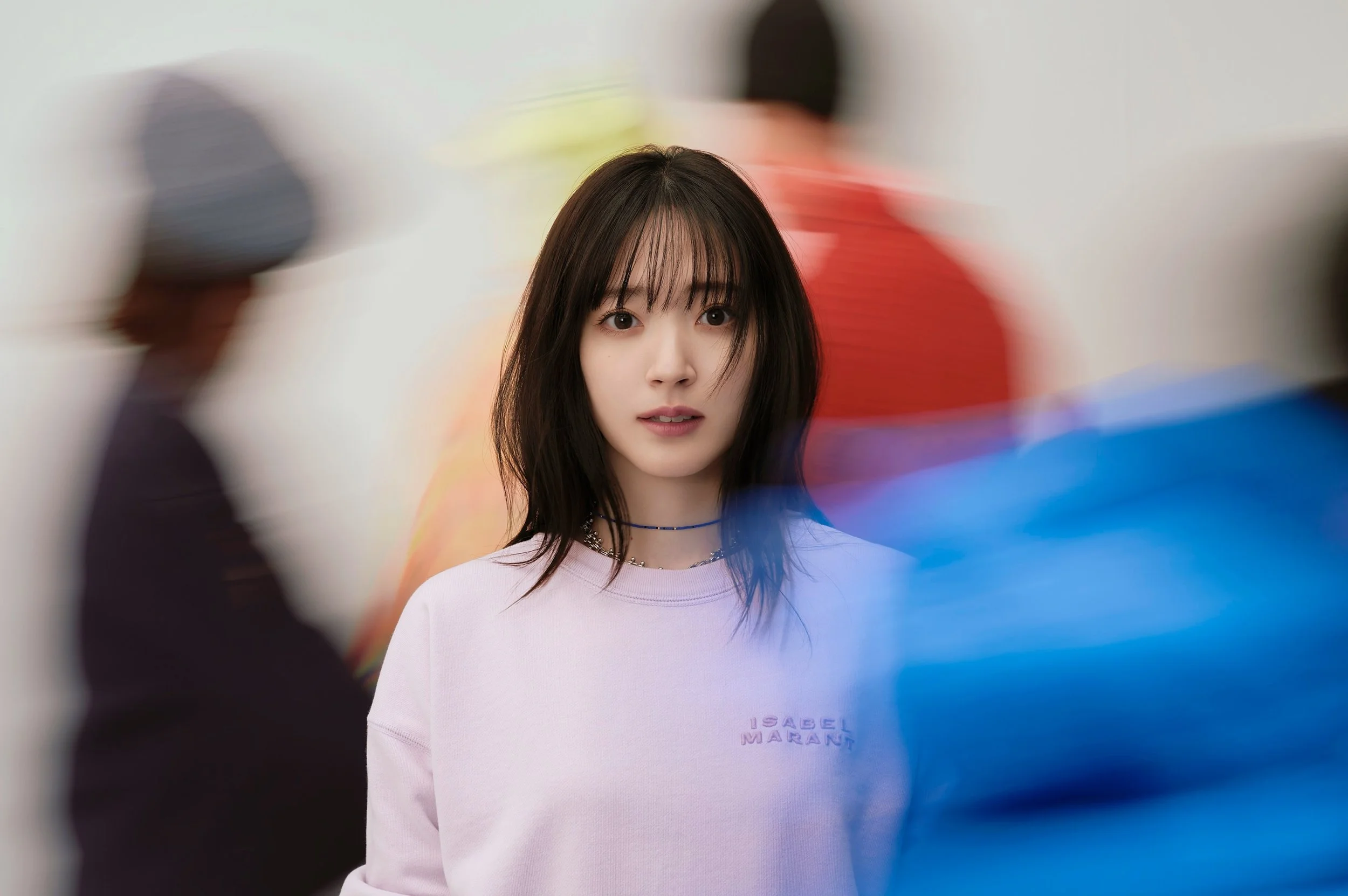 しゃがんでいる若い女性が中心に写り、周囲には色とりどりの布や服を着た人々が動きながら隣接している。女性は白い長袖のトップスとネックレスを着用している。