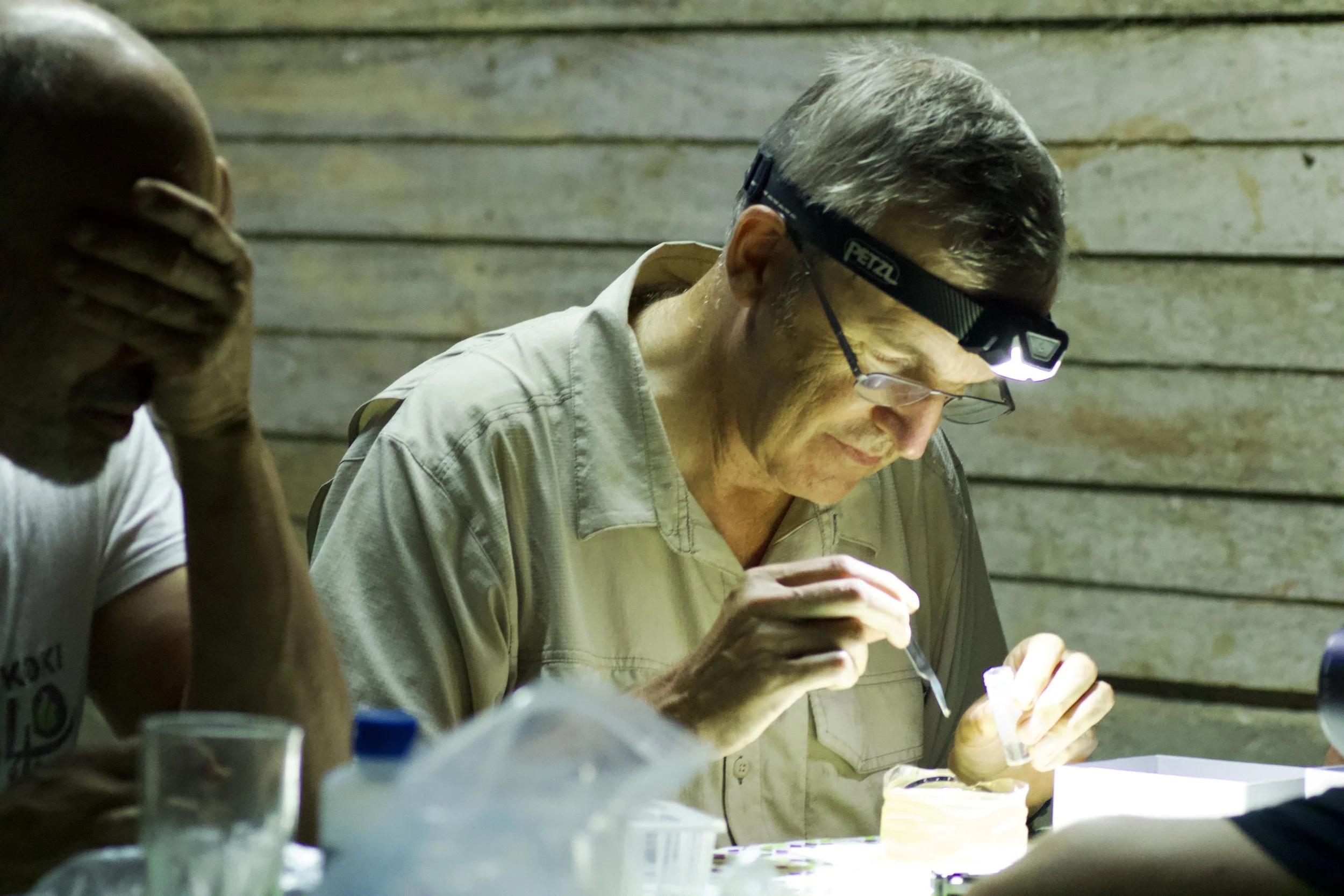 Une personne portant des lunettes frontales éclairant une surface, avec une autre personne à gauche, dans un environnement en bois.